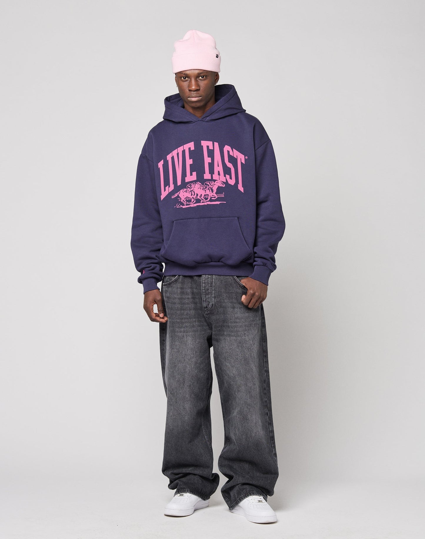 A person stands against a plain gray background wearing the LIVE FAST DIE YOUNG Live Fast Stallion Hooded, an oversized navy hoodie with a bold pink LIVE FAST front print, paired with a light pink beanie, baggy dark jeans, and white sneakers.