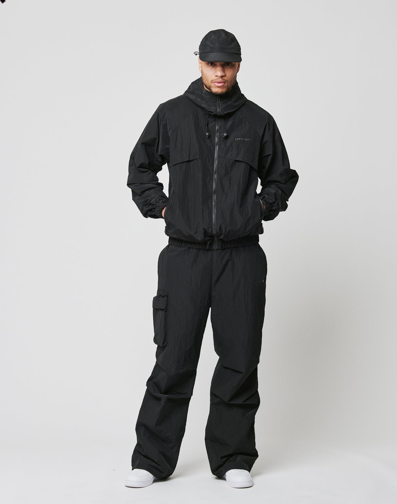 Against a plain white background, a person wears an oversized black zip-up jacket and LIVE FAST DIE YOUNG Live Fast Nylon Pants with an elastic waistband, paired with white sneakers. Their hands are in their jacket pockets.
