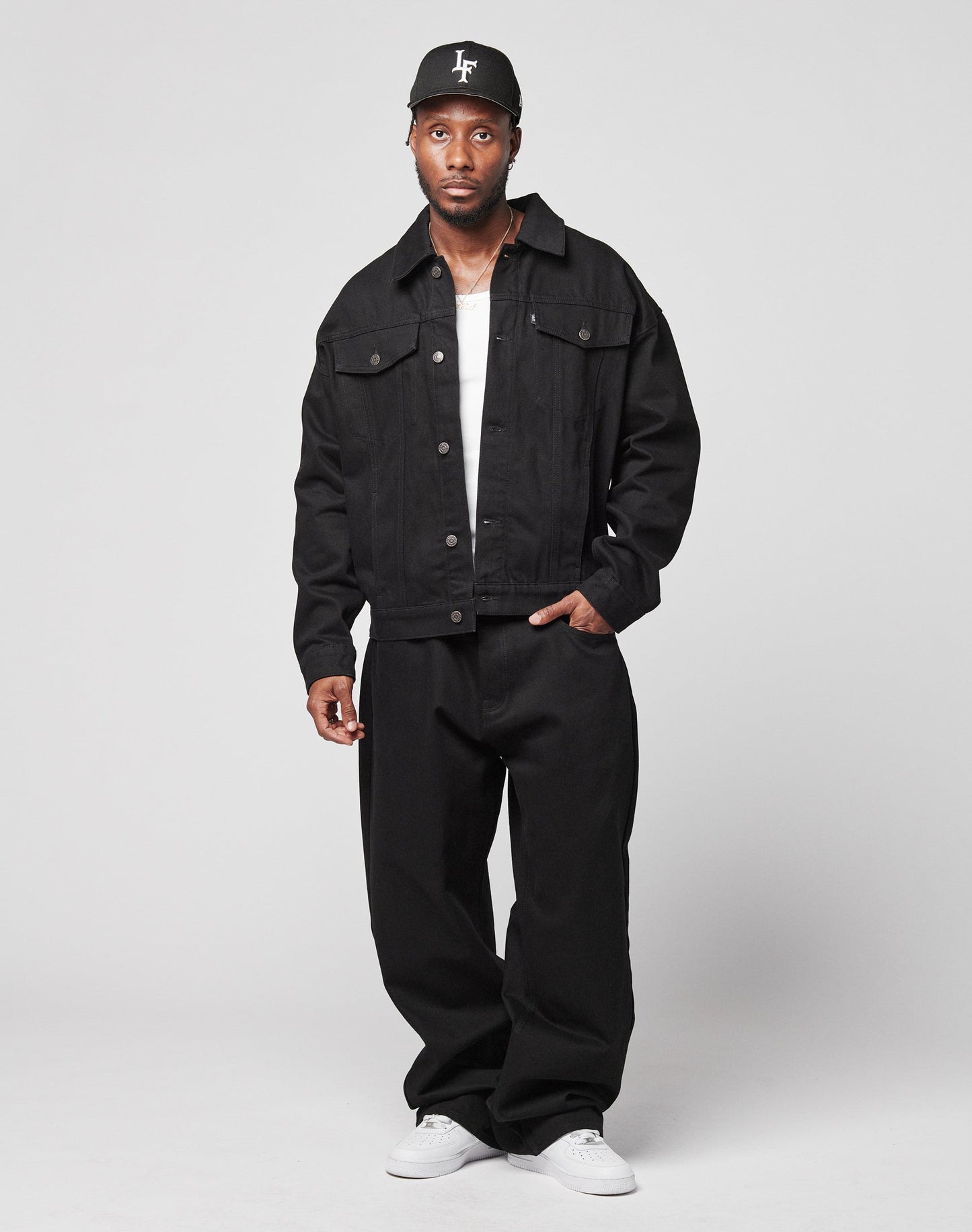 A man stands against a plain background wearing the LFDY Denim Jacket by LIVE FAST DIE YOUNG, loose black pants, a white shirt, white sneakers, and a black baseball cap with white lettering. He looks directly at the camera with a neutral expression.