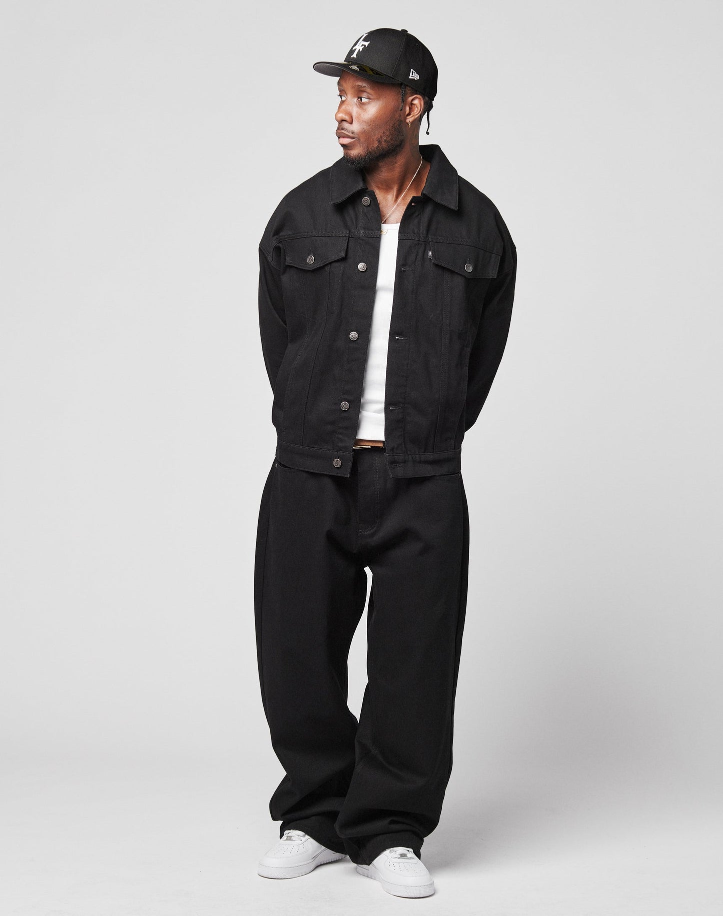 A man stands against a plain background wearing the LIVE FAST DIE YOUNG LFDY Baggy Denim jacket, wide-leg black pants, a white shirt, white sneakers, and a black baseball cap. His hands are behind his back as he looks to the side.