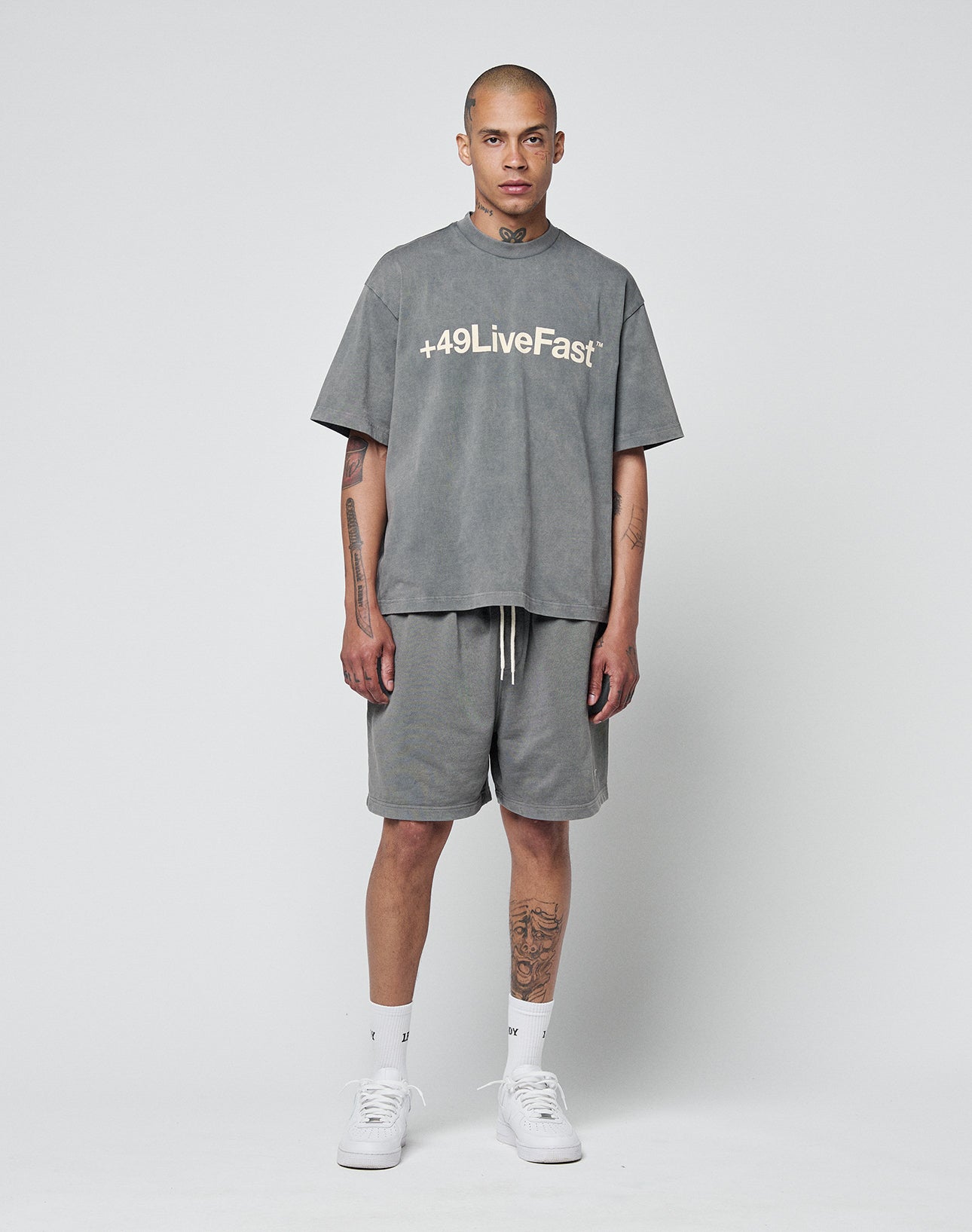 A person wearing the LIVE FAST DIE YOUNG Basic +49 Tee, an oversize 100% cotton grey T-shirt with “+49LiveFast” print, pairs it with grey shorts, white socks, and sneakers. Tattoos are visible on their arms and neck.