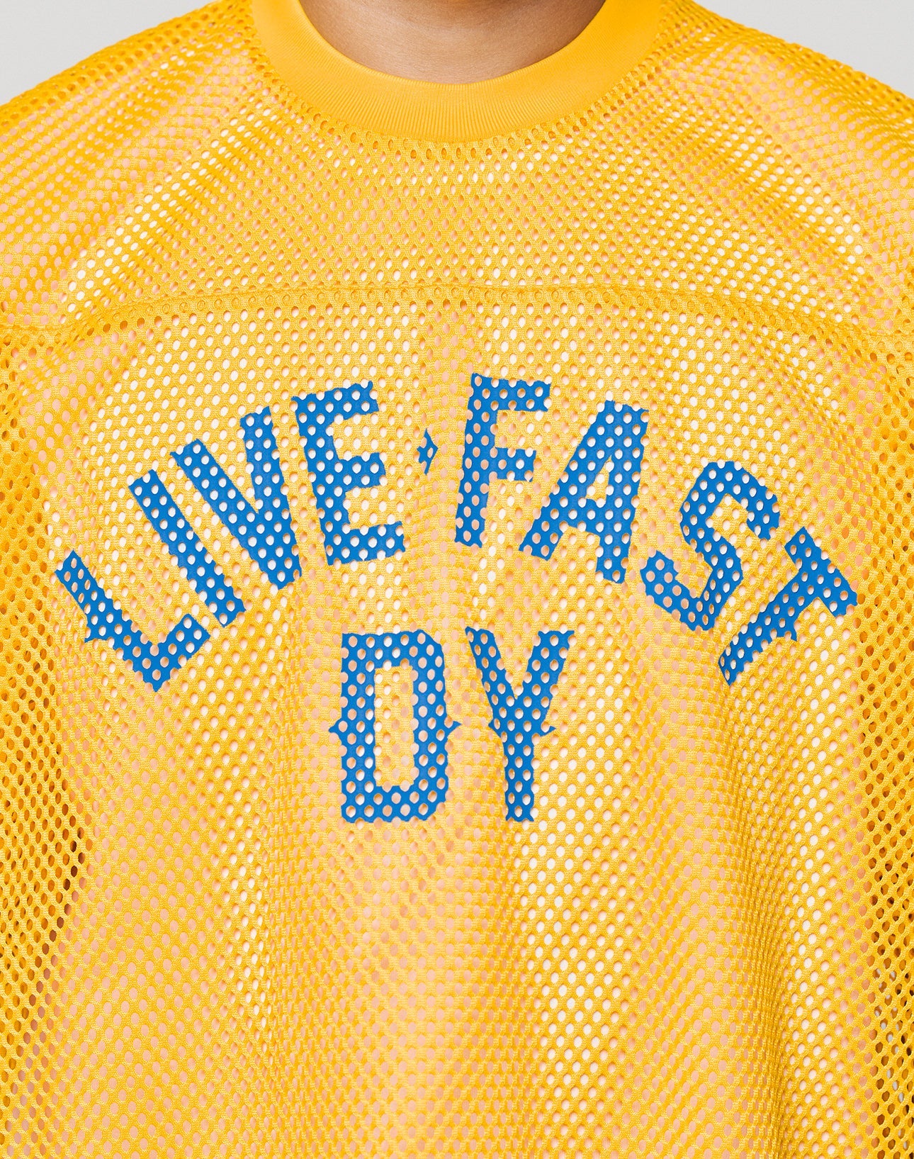 A person wears the LFDY Boxy Jersey by LIVE FAST DIE YOUNG, a yellow mesh shirt with blue LIVE FAST DY lettering, yellow collar, boxy fit, and a partially obscured front print. The shirt is shown from shoulders to upper chest.