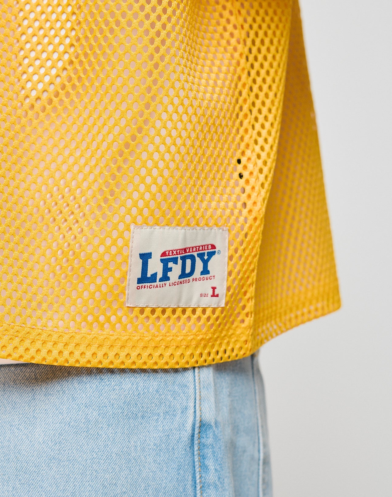 Close-up of a person wearing the LFDY Boxy Jersey by LIVE FAST DIE YOUNG—a yellow mesh top with boxy fit, paired with light blue jeans. The white tag reads LFDY OFFICIALLY LICENSED PRODUCT, size L in red.
