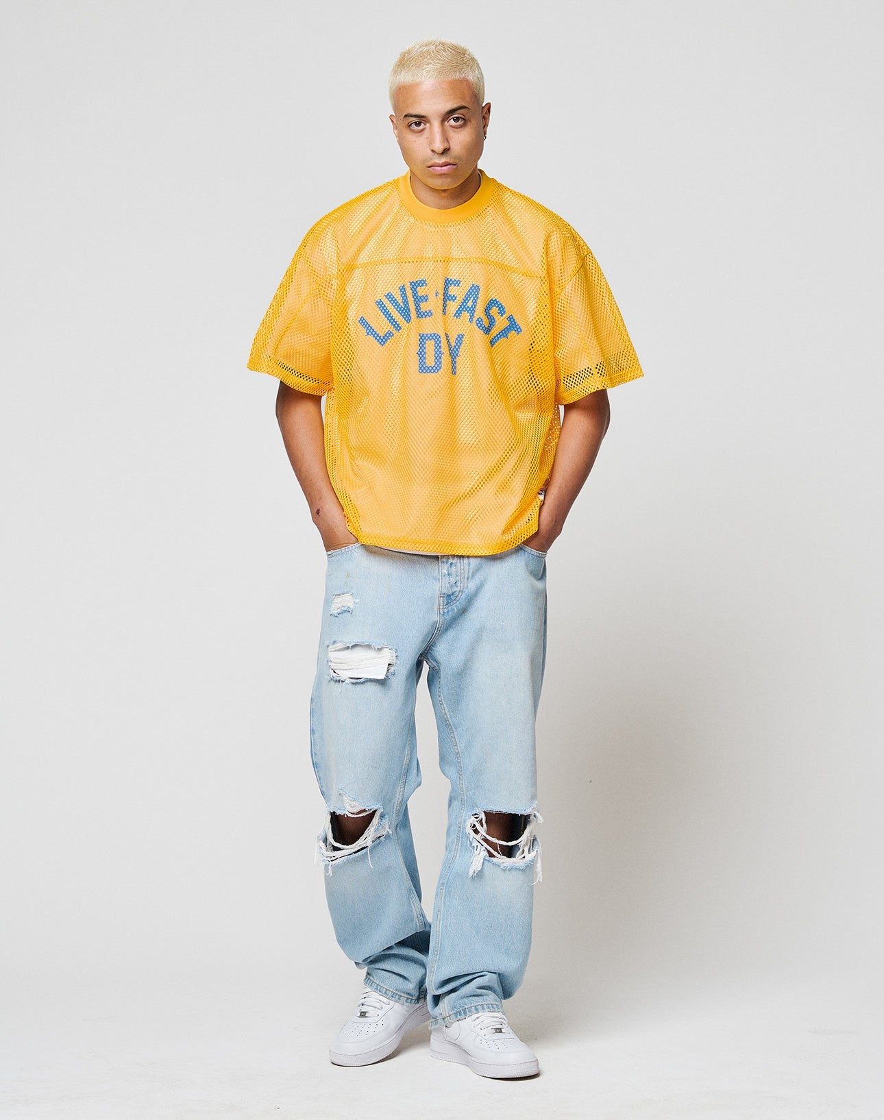 A person with short blond hair wears the LFDY Boxy Jersey by LIVE FAST DIE YOUNG, paired with light blue ripped jeans and white sneakers, standing against a plain white background.
