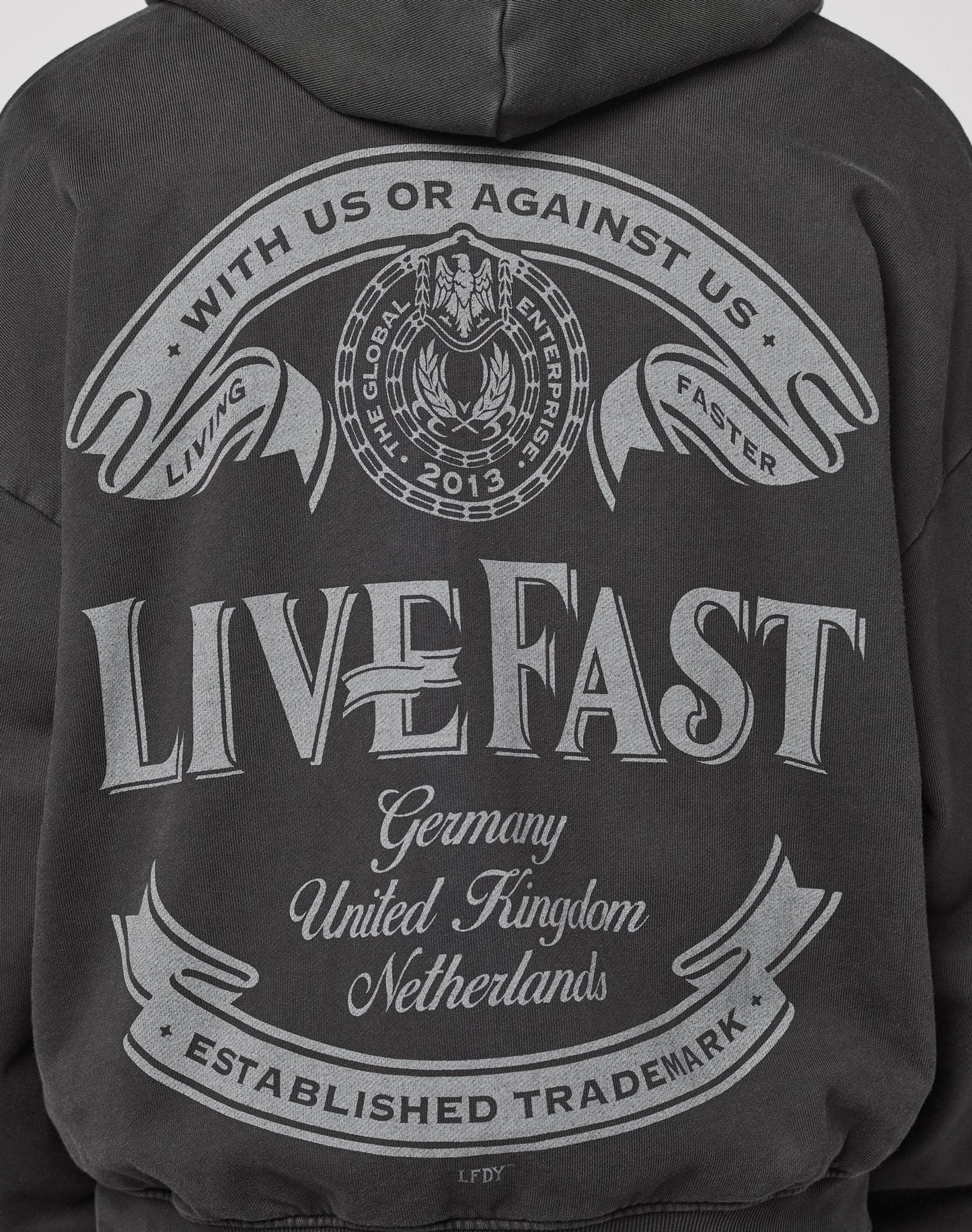 The LFDY Loyal Hooded Zip by LIVE FAST DIE YOUNG is an oversized, dark hoodie in 100% cotton with a vintage wash and bold back graphic—WITH US OR AGAINST US, LIVING FAST—with decorative banners listing Germany, UK, Netherlands.