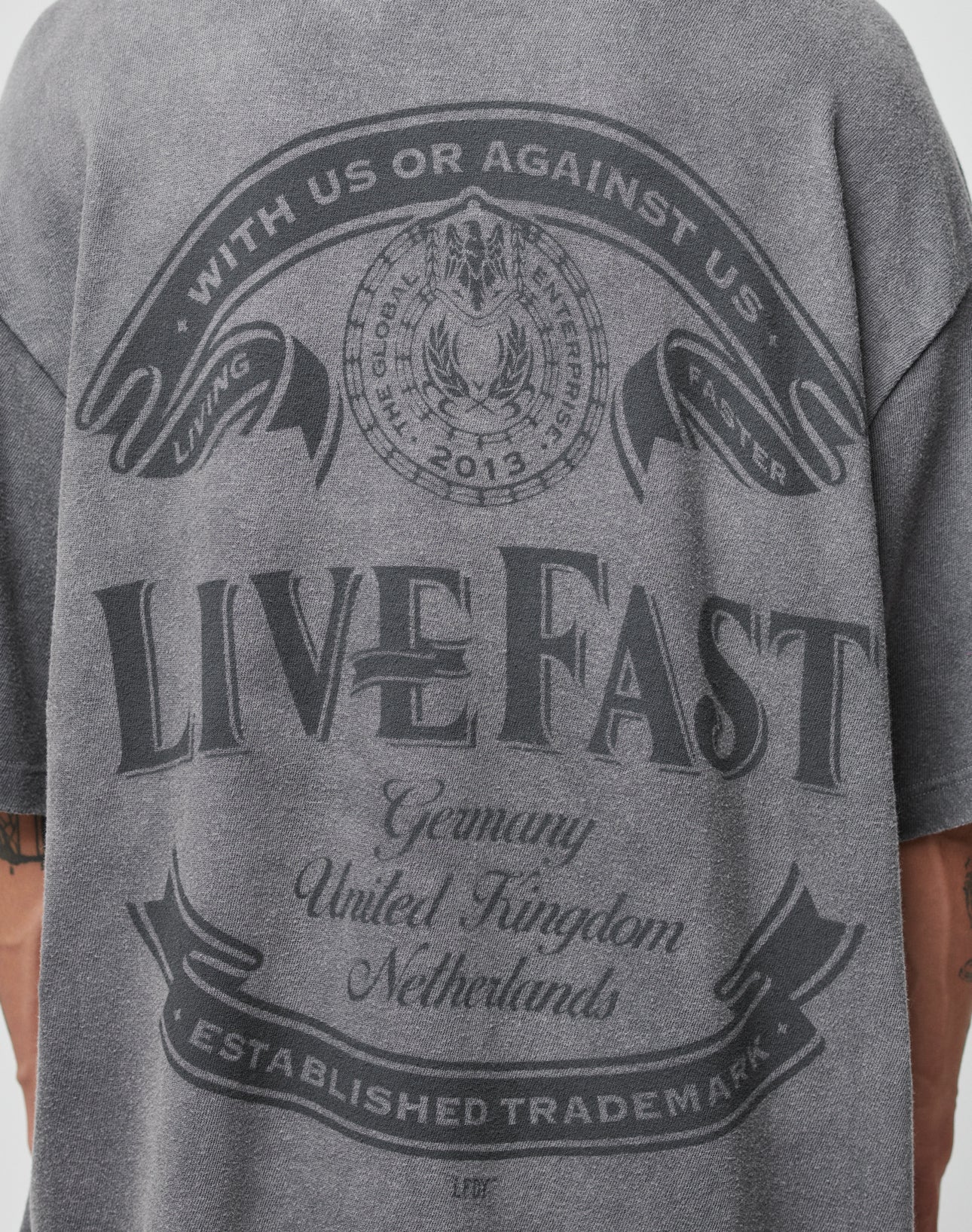 A person wears a gray LFDY Loyal Tee by LIVE FAST DIE YOUNG featuring “With us or against us. Living faster. The Good Life Established 2012. Live Fast.” Oversize fit, 100% Baumwolle, with Germany, UK, and Netherlands listed.