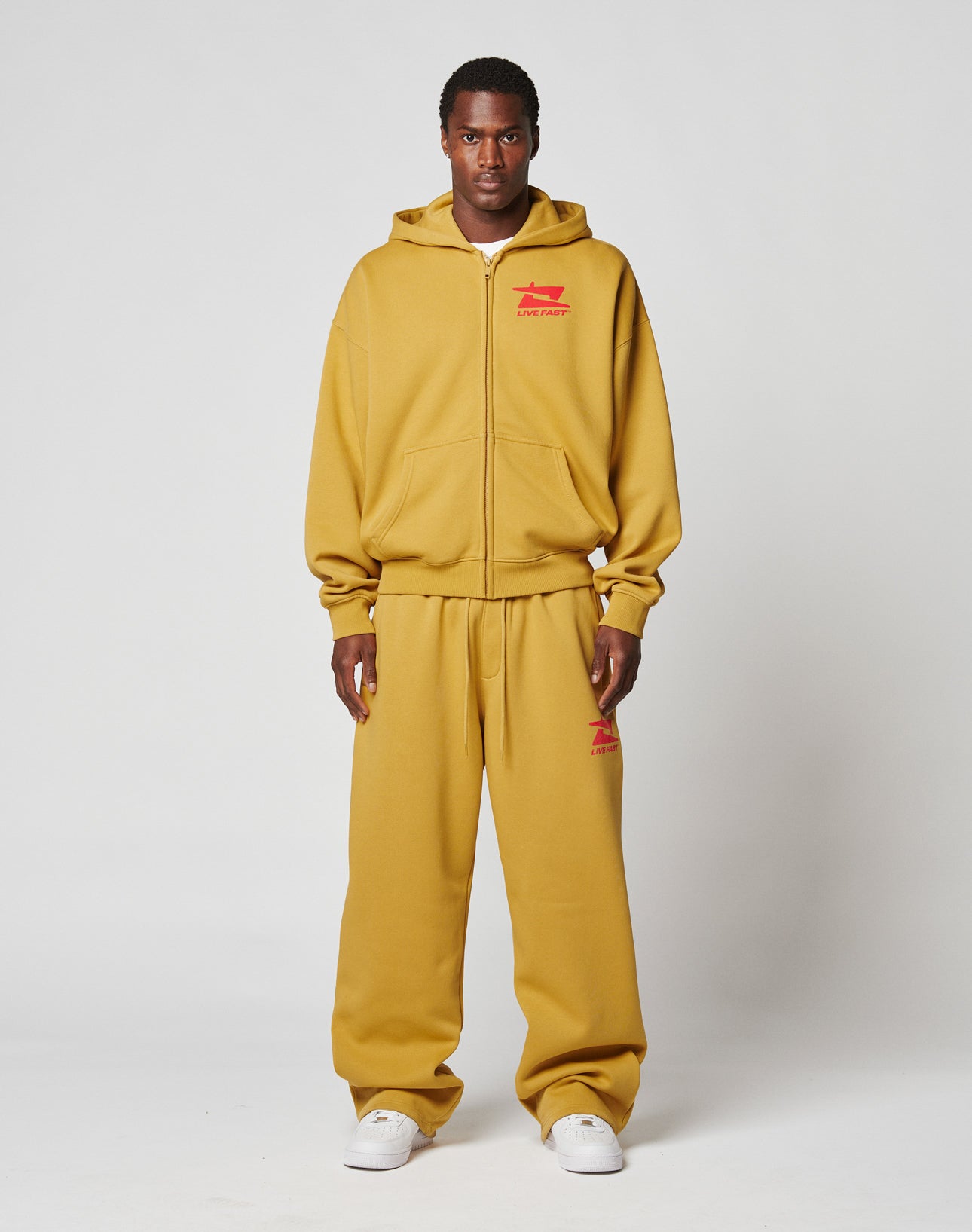 A person faces forward in a mustard yellow LIVE FAST DIE YOUNG Live Fast Sweatsuit Jacket with matching red frontprint sweats and white sneakers, standing against a plain light background.