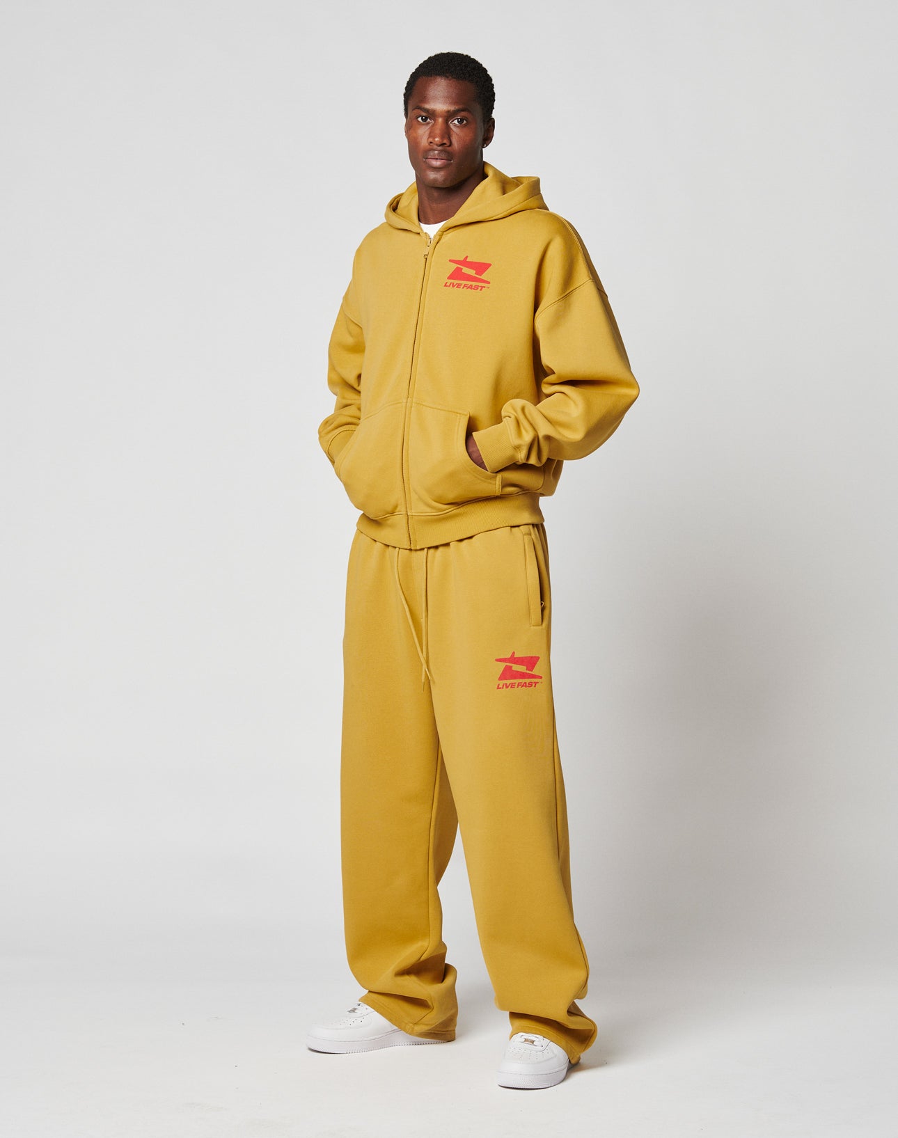 A man stands against a white background wearing the LIVE FAST DIE YOUNG Live Fast Sweatsuit Jogger in mustard yellow with red graphics, an elastik-bund, and white sneakers.