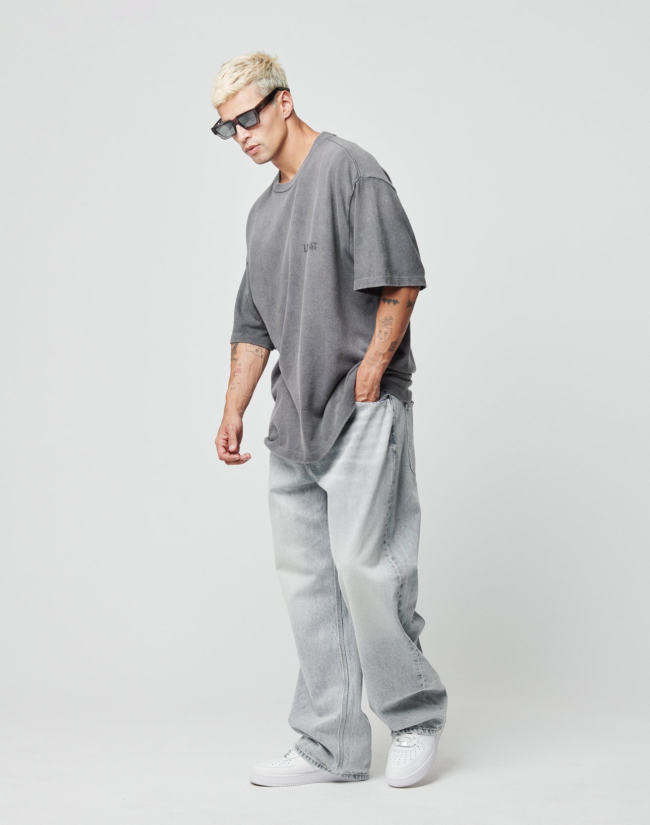 A man with short blond hair wearing black sunglasses, a loose gray t-shirt, and LIVE FAST DIE YOUNG LFDY Baggy Denim (weite gerades Bein, light-wash, 100% Baumwolle) stands against a white background with white sneakers.
