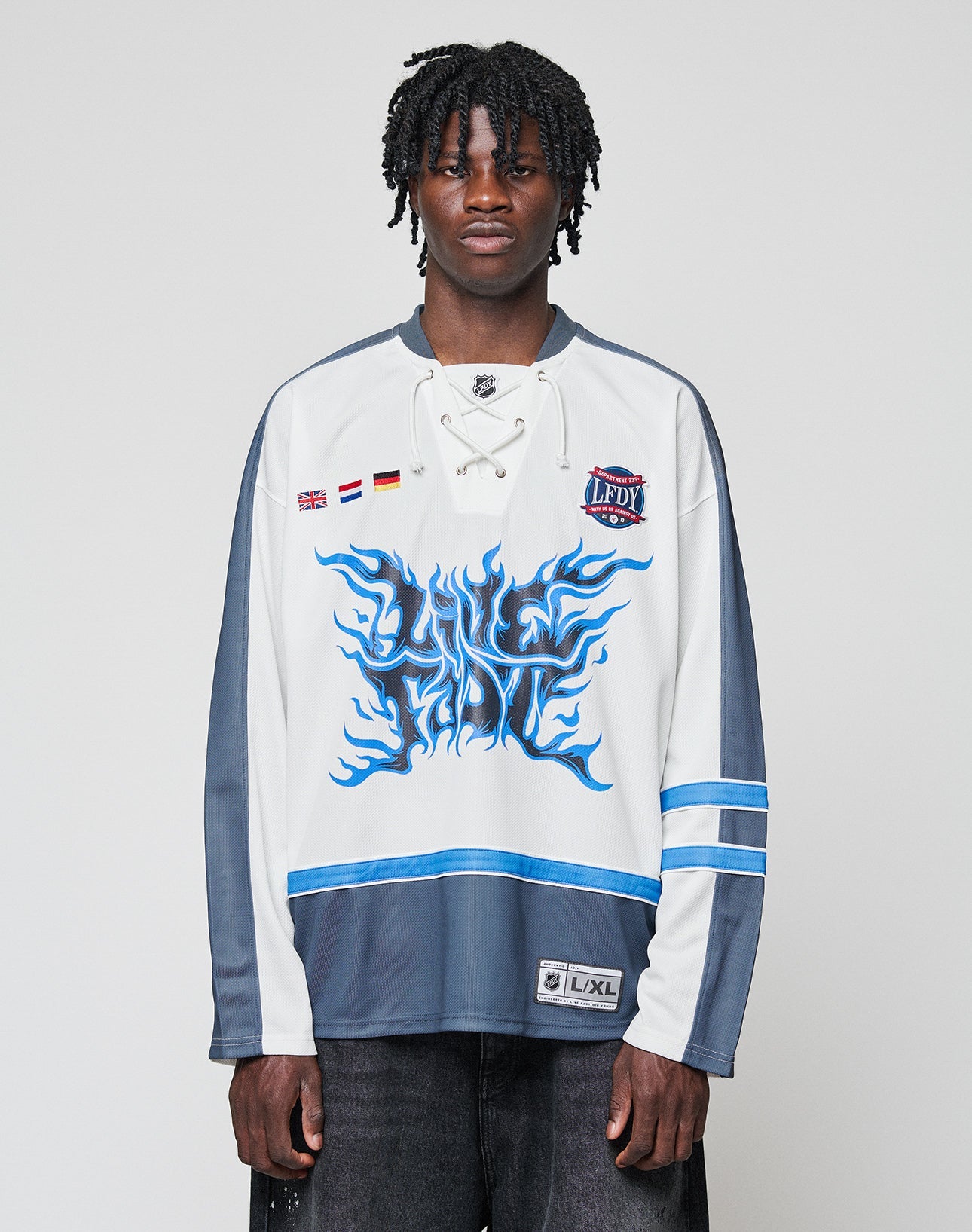 A man with short dreadlocks wears the LIVE FAST DIE YOUNG LFDY Flames Jersey—an oversized white and gray hockey top with blue flame graphics, front print details, and stripe patches—standing against a plain light background.