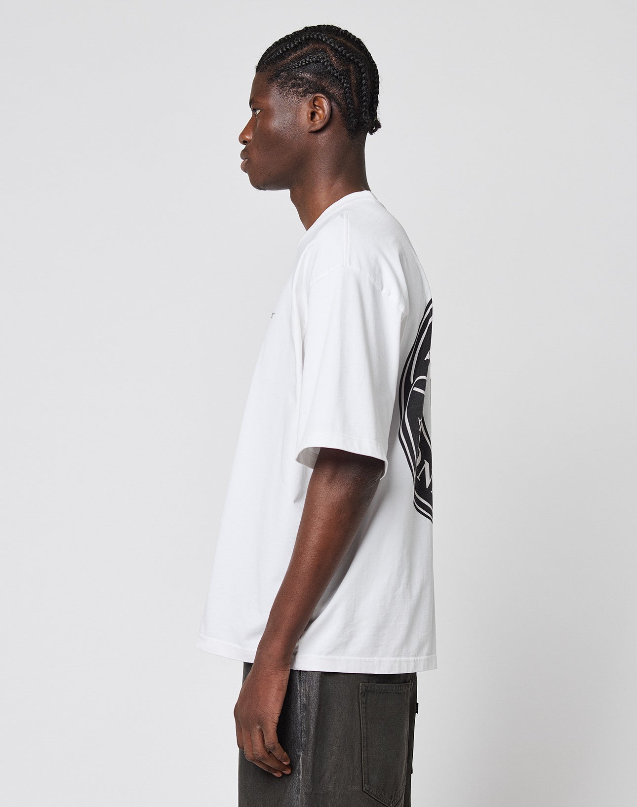 A man with braided hair is seen in profile wearing the LIVE FAST DIE YOUNG LFDY Monaco Boys Tee, an oversize-fit white T-shirt with a large black back graphic made from 100% Baumwolle, paired with dark pants against a plain light background.