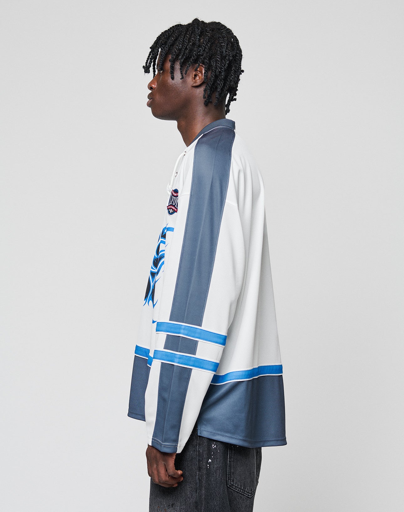 A person with short, twisted hair stands in profile, wearing the LIVE FAST DIE YOUNG LFDY Flames Jersey—an oversize white and gray hockey-style top with blue accents and a bold front print—paired with black pants against a plain light background.