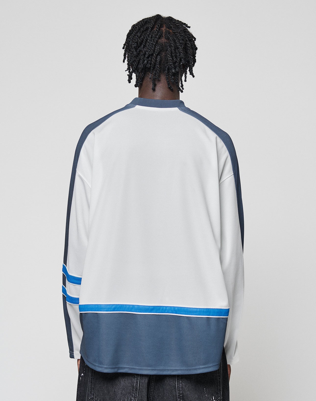 A person with short, twisted hair is shown from the back, wearing the LIVE FAST DIE YOUNG LFDY Flames Jersey—an oversized long-sleeve white and gray shirt with blue accents—and black pants against a plain light background.