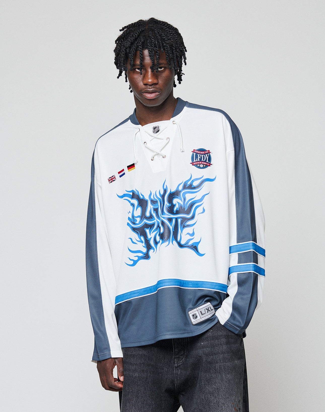 A person with short, twisted hair wears the LIVE FAST DIE YOUNG LFDY Flames Jersey—a blue flame print, oversized polyester hockey-style top with patches—standing against a plain light gray background.