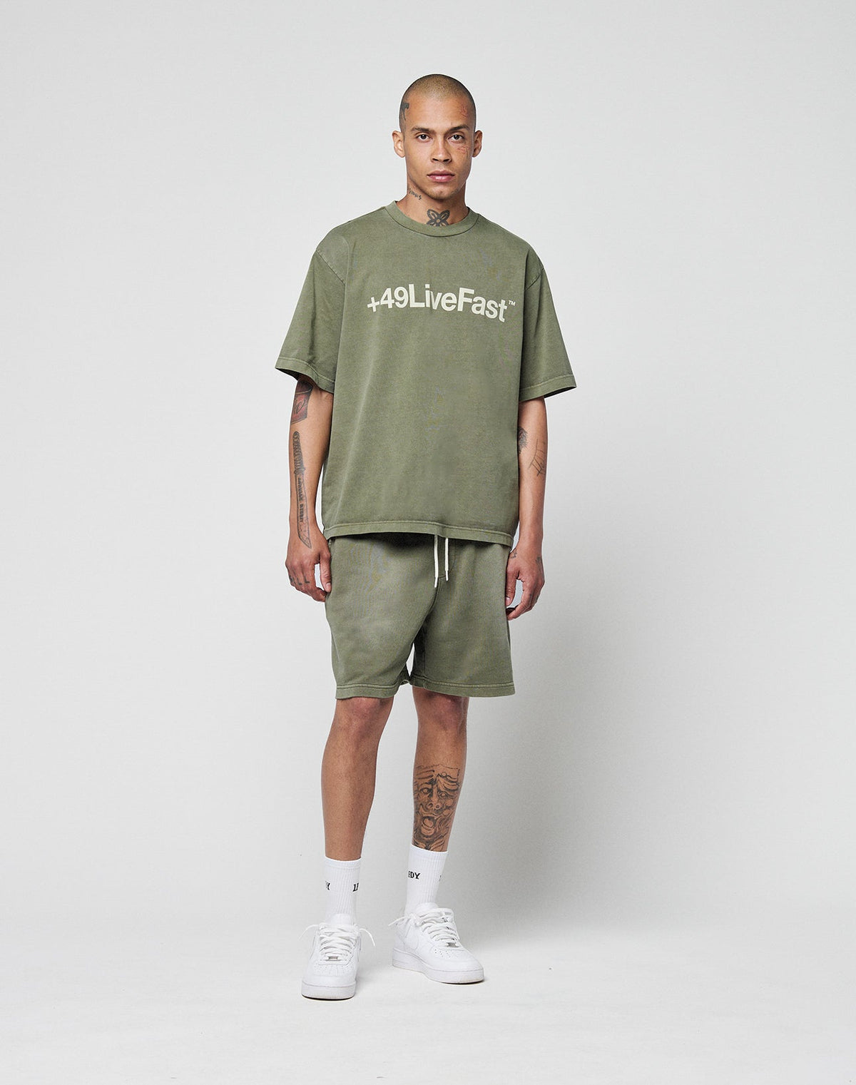 A tattooed person with a neutral expression stands against a white background, wearing olive green LIVE FAST DIE YOUNG Basic Sweat Shorts with an elastic waistband, an olive “+49LiveFast” tee, white socks, and white sneakers.