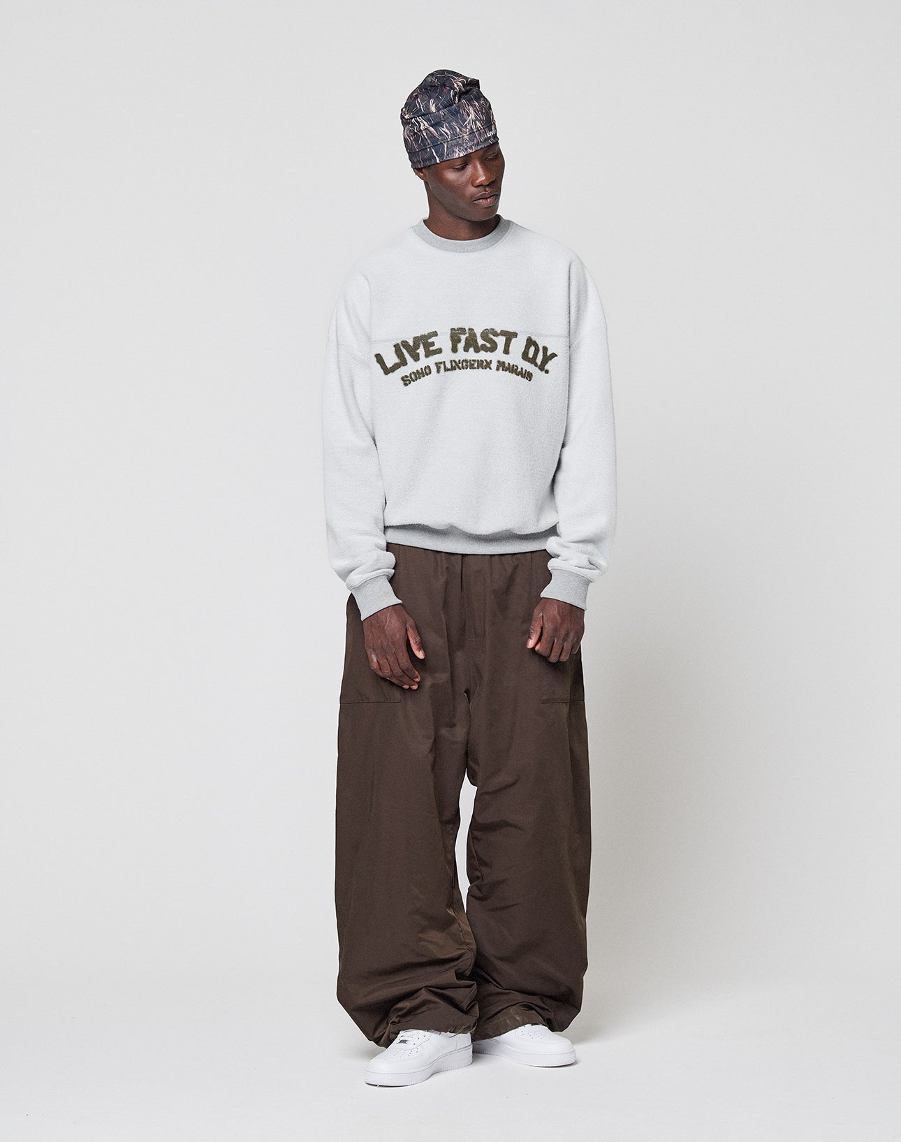 A man stands against a plain background wearing a patterned headscarf, a live fast die sweatshirt, LIVE FAST DIE YOUNG Baggy Nylon Pants in brown with an elastic waistband, and white sneakers. He looks to the side with his hands relaxed.