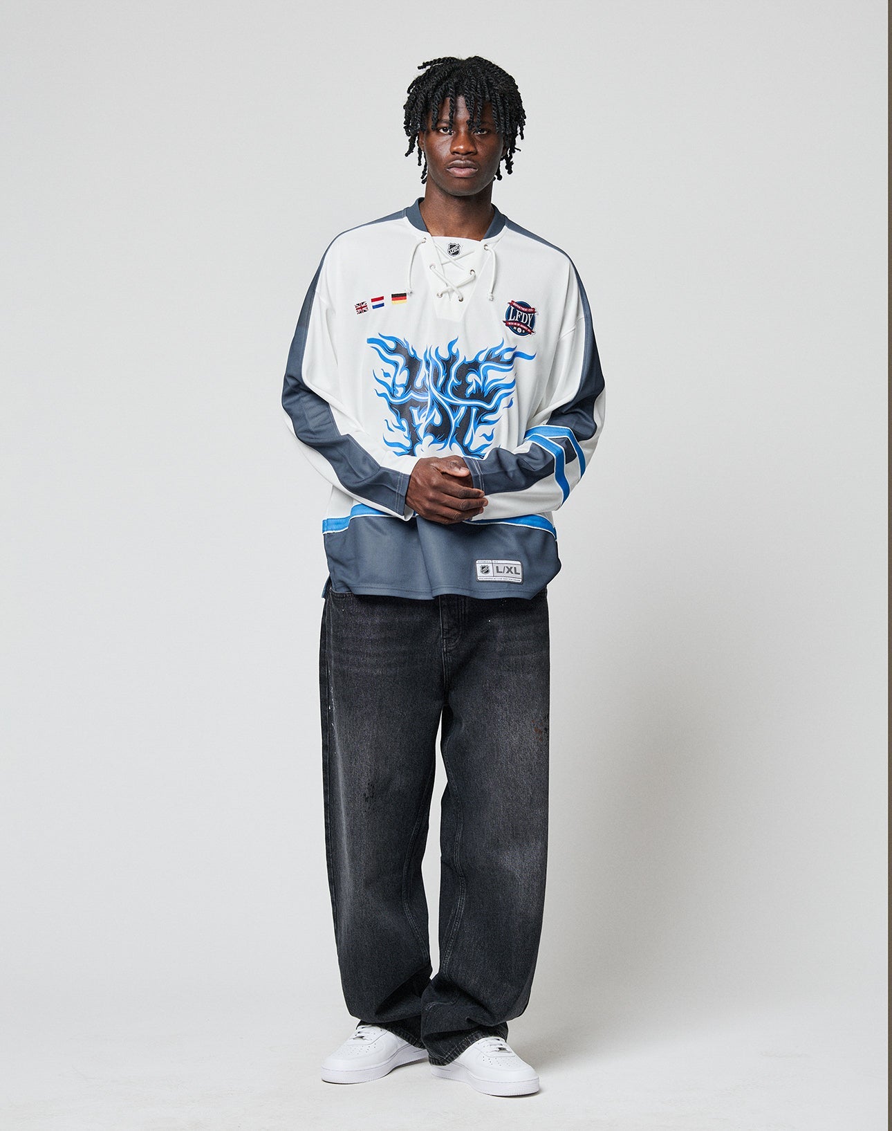 A person with short dreadlocks stands against a plain background, wearing the LIVE FAST DIE YOUNG LFDY Flames Jersey—an oversize polyester hockey-style top with a blue flame frontprint—paired with loose black jeans and white sneakers, hands clasped in front.