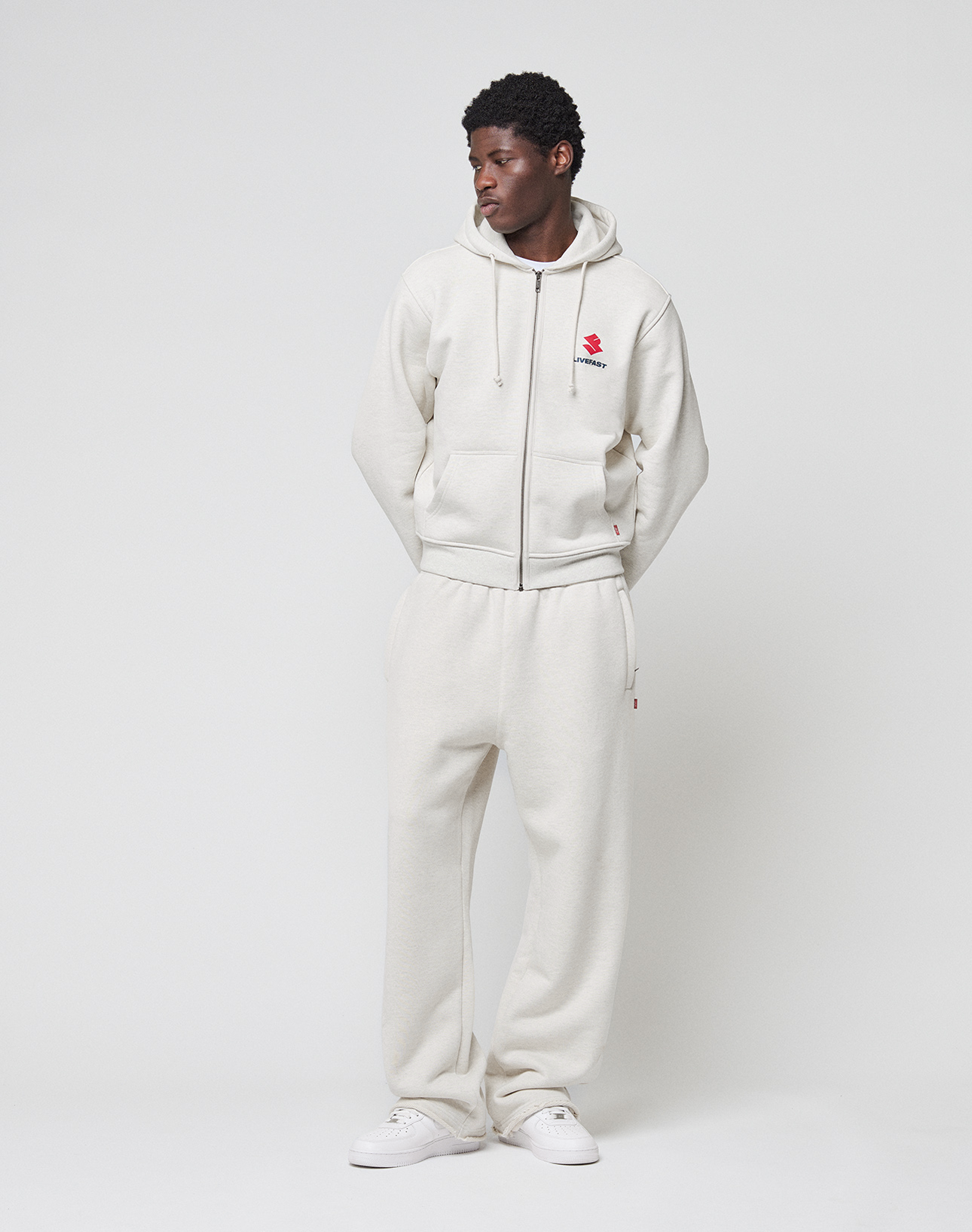 A man stands against a white background wearing the LIVE FAST DIE YOUNG LFDY Heavyweight Lounge Zip Hooded in light beige and matching sweatpants with white sneakers. His hands are in his pockets as he looks slightly to the side, highlighting the regular fit.