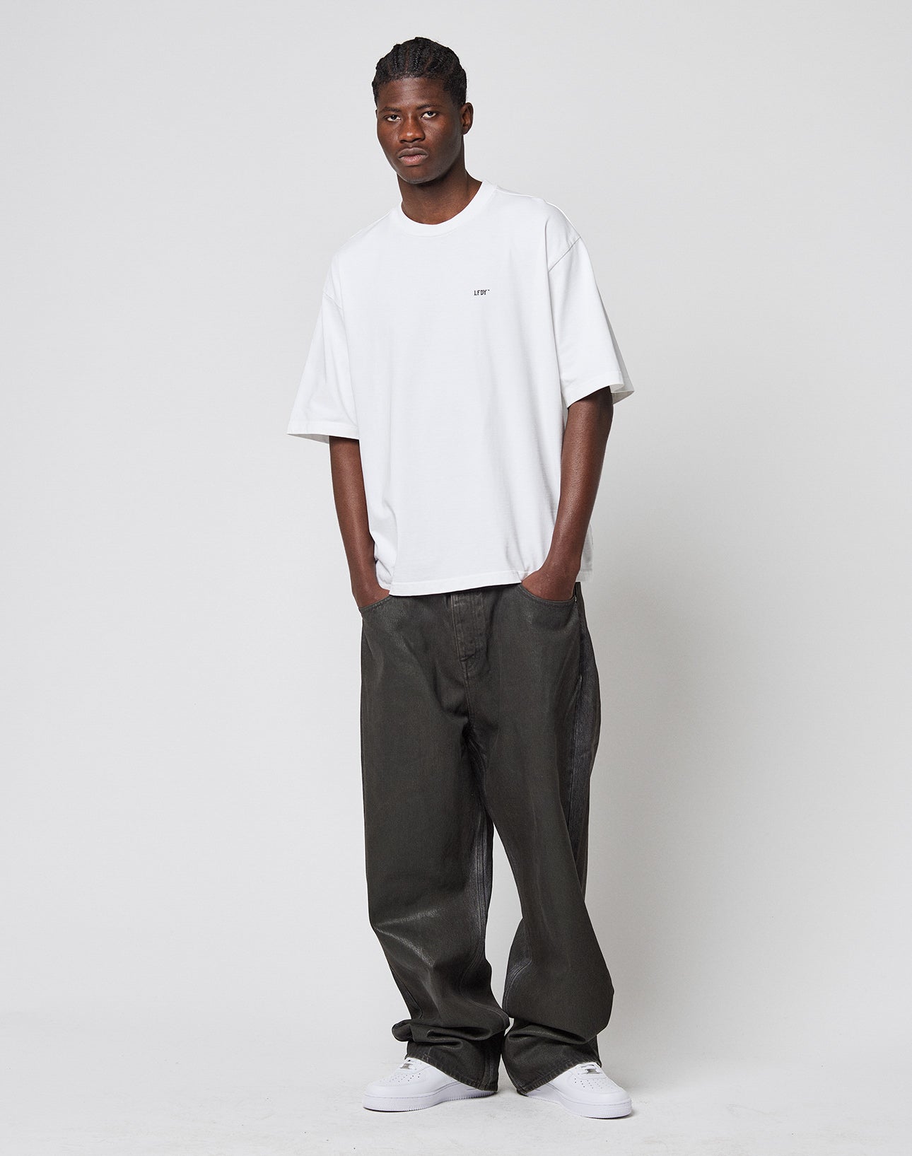 A man stands against a plain white background wearing the LIVE FAST DIE YOUNG LFDY Monaco Boys Tee in white, paired with oversized dark gray vintage-look pants and white sneakers, hands in pockets as he looks calmly at the camera.