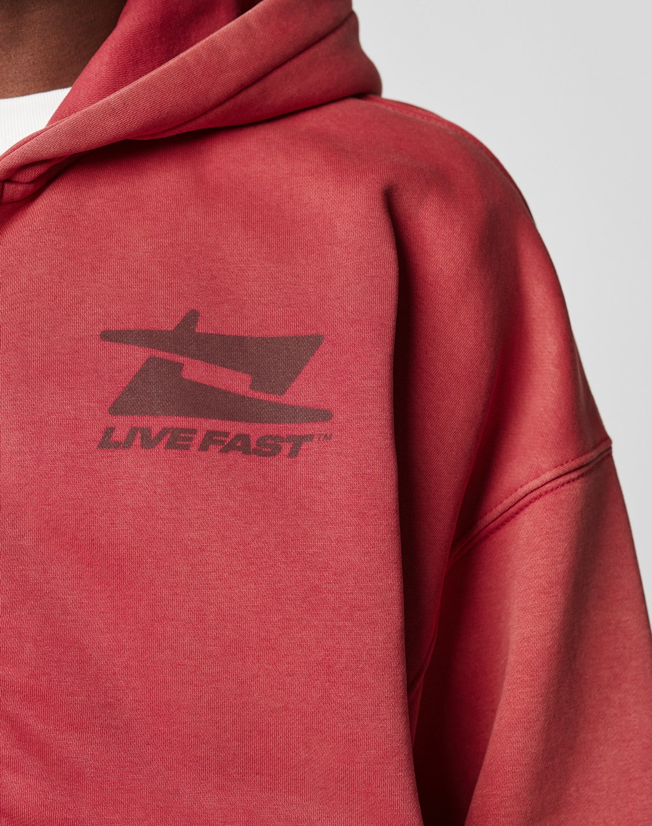 A person wears the LIVE FAST DIE YOUNG Live Fast Sweatsuit Jacket in faded red with a vintage wash, featuring an airplane logo and LIVE FAST text on the chest. The hood is up, showing an oversize fit and a white shirt underneath.