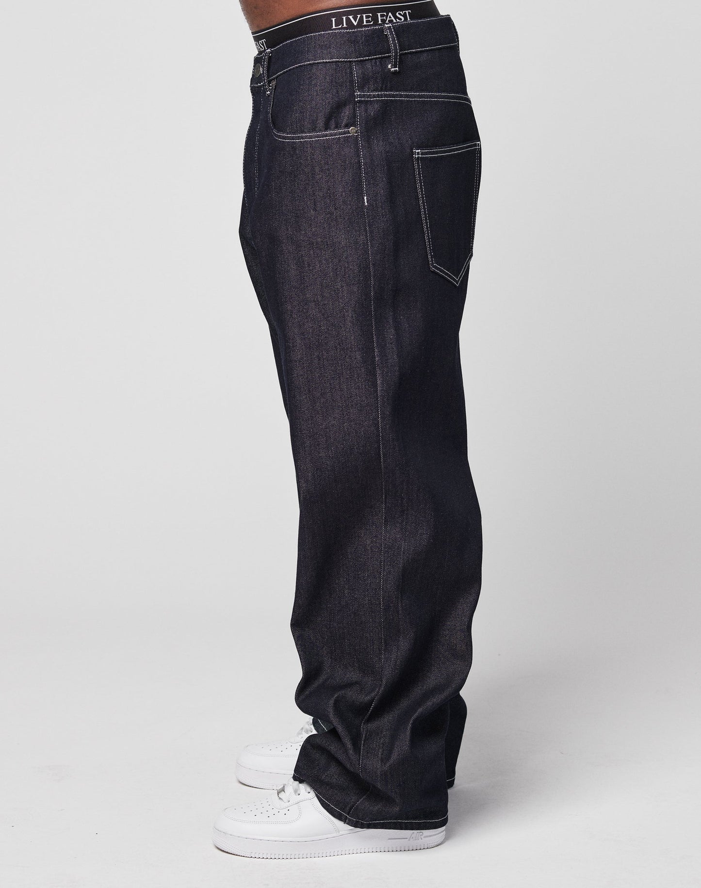 A person wearing dark blue LFDY Baggy Denim jeans with a LIVE FAST waistband and white sneakers, standing sideways against a plain light background.