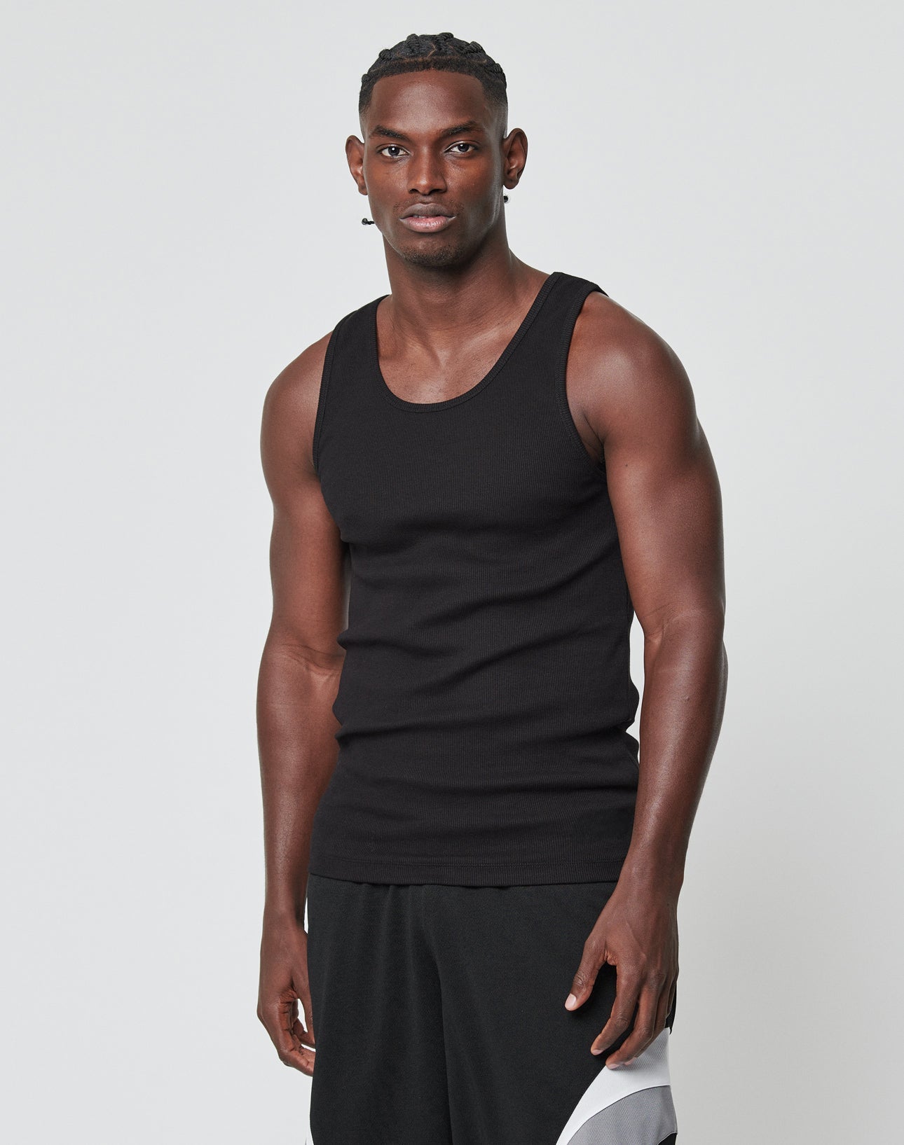 A man with short braids wears the LIVE FAST DIE YOUNG Blank Rib Tank and black shorts featuring a white design and flag label, standing against a plain light background and looking at the camera with a neutral expression.
