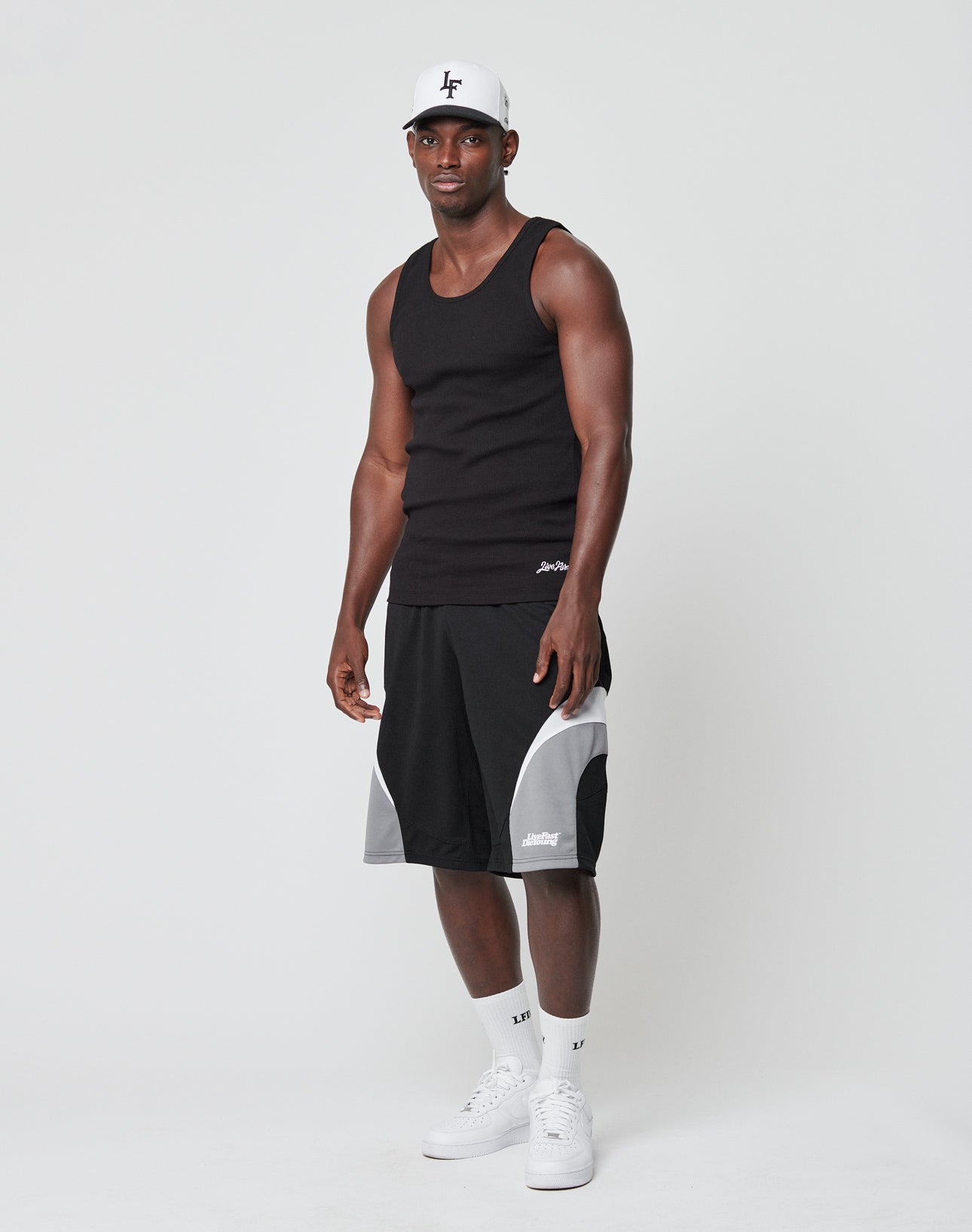 A man in a LIVE FAST DIE YOUNG Basic Rib Tank, black and gray athletic shorts, white crew socks, white cap, and white sneakers stands against a plain white background.