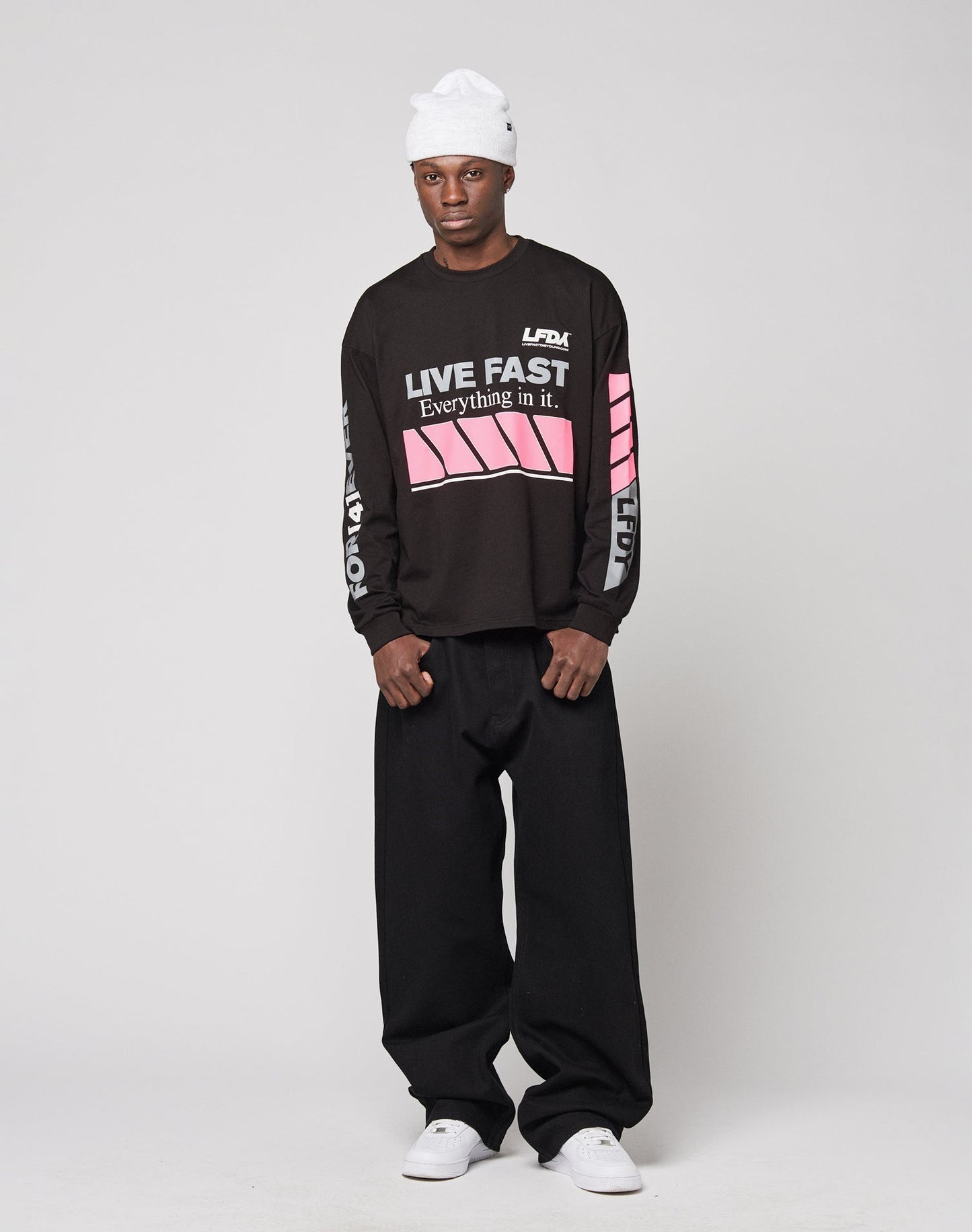 A man wears an oversize black LFDY Racing Longsleeve by LIVE FAST DIE YOUNG with pink reflective print, paired with loose black pants, a white beanie, and white sneakers against a plain light background.