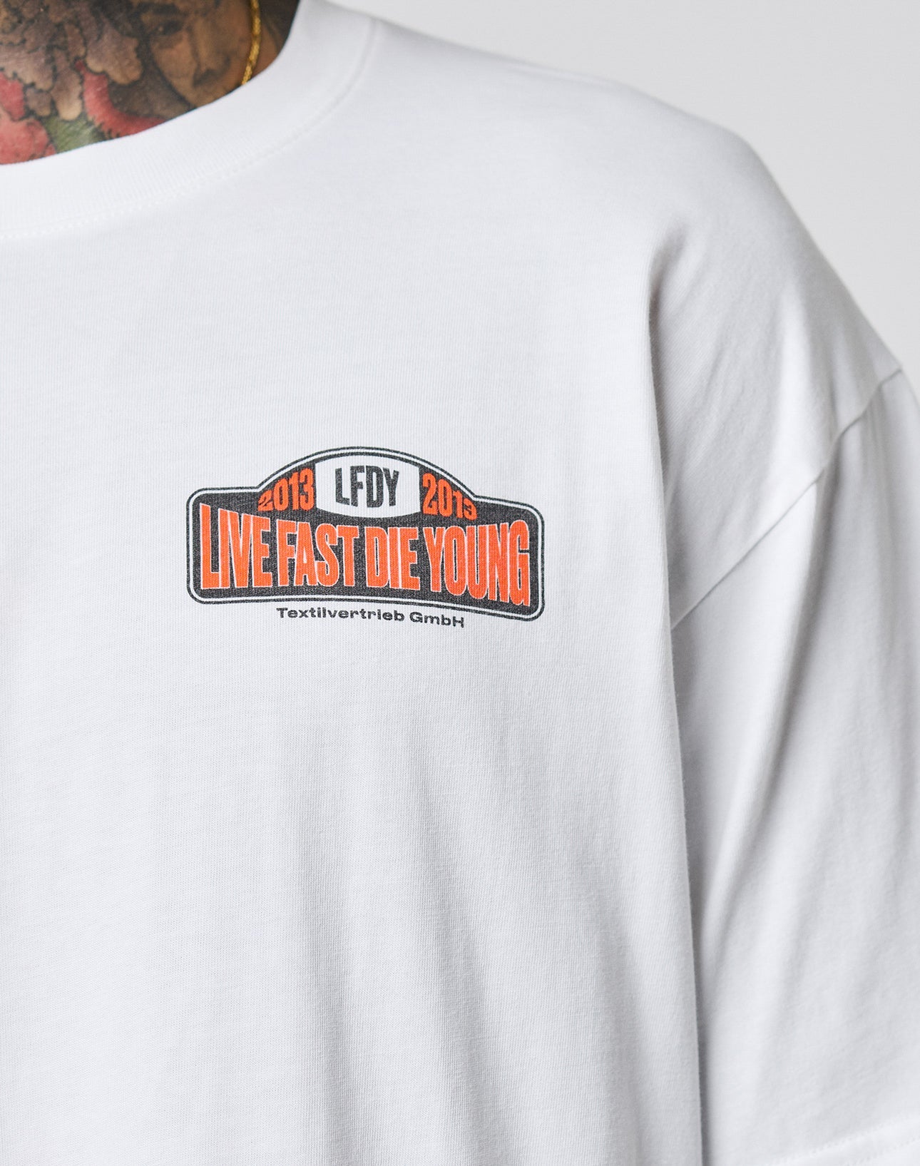 A person in the Live Fast 2013 Tee by LIVE FAST DIE YOUNG—a regular fit white shirt with LIVE FAST DIE YOUNG in bold orange, 2013 LFDY 2013, and Textilvertretieb GmbH on a chest patch. Part of a tattooed neck is visible.