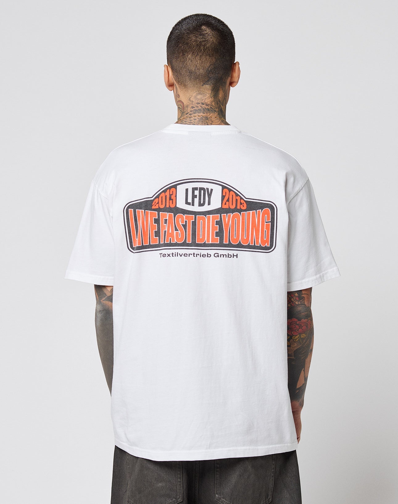 A tattooed individual wears the Live Fast 2013 Tee by LIVE FAST DIE YOUNG, a white Regular Fit t-shirt made of 100% cotton, featuring bold back prints and clean topstitching.