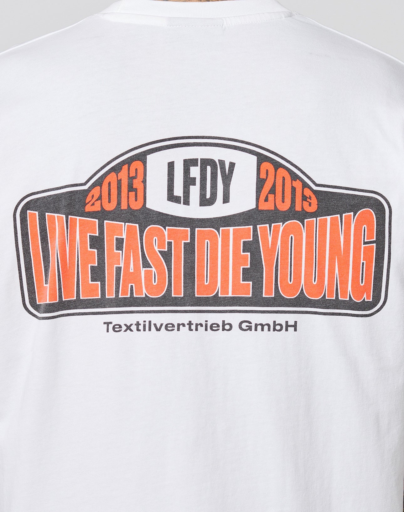 The Live Fast 2013 Tee by LIVE FAST DIE YOUNG is a white regular fit t-shirt made from 100% Baumwolle, featuring “LFDY” and “2013 2019” above bold orange and black “LIVE FAST DIE YOUNG” text with “Textilvertrieb GmbH” below.