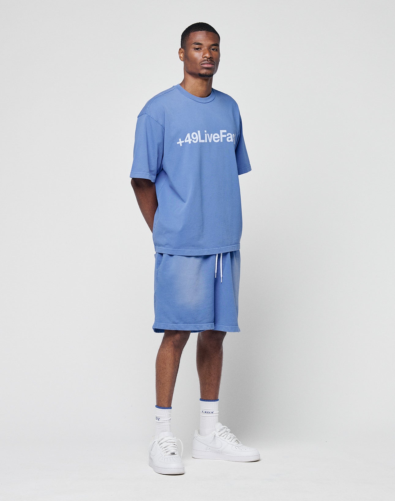 A man stands against a white background wearing the Basic +49 Tee by LIVE FAST DIE YOUNG in light blue, with +49LiveFair printed on the front, matching blue shorts, white socks, and white sneakers. His arms are behind his back.
