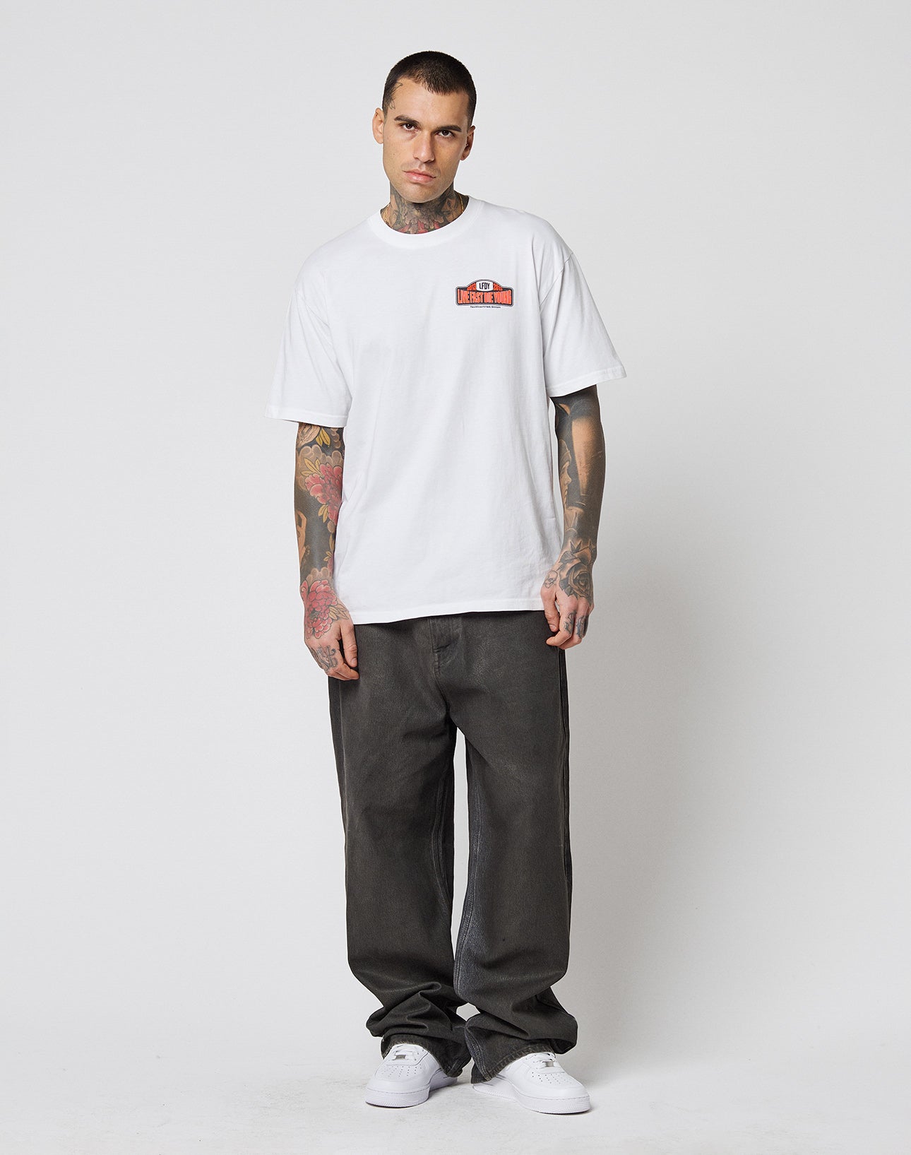 A tattooed man stands against a white background, wearing the LIVE FAST DIE YOUNG Live Fast 2013 Tee (100% Baumwolle, small red chest graphic), baggy dark gray jeans with topstitching, and white sneakers. He looks directly at the camera.