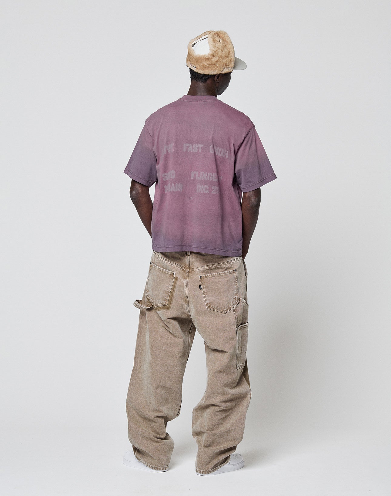 A person stands facing away, wearing the LIVE FAST DIE YOUNG Live Fast District Tee in faded purple, paired with baggy light brown jeans with exposed pockets, white sneakers, and a tan and white furry cap against a plain white background.