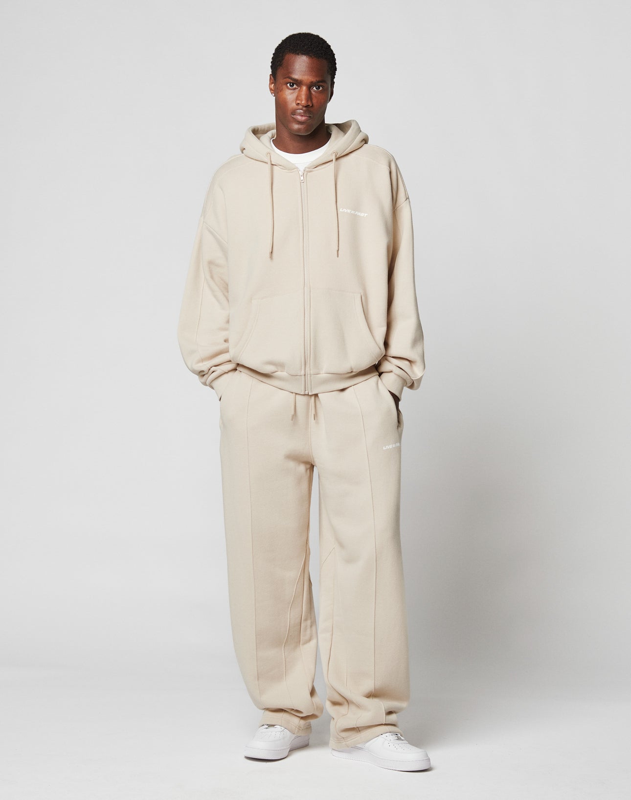 A man stands facing forward, wearing a LIVE FAST DIE YOUNG LFDY Pleated Hooded Zip in beige with matching sweatpants and white sneakers, set against a plain light grey background.