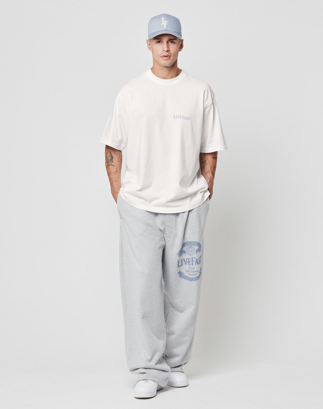 A man stands against a plain background wearing a light blue cap, Oversize Fit white T-shirt, white sneakers, and LIVE FAST DIE YOUNG Loyal Wide Jogger sweatpants with elastik-bund and logo, looking at the camera with hands in pockets.