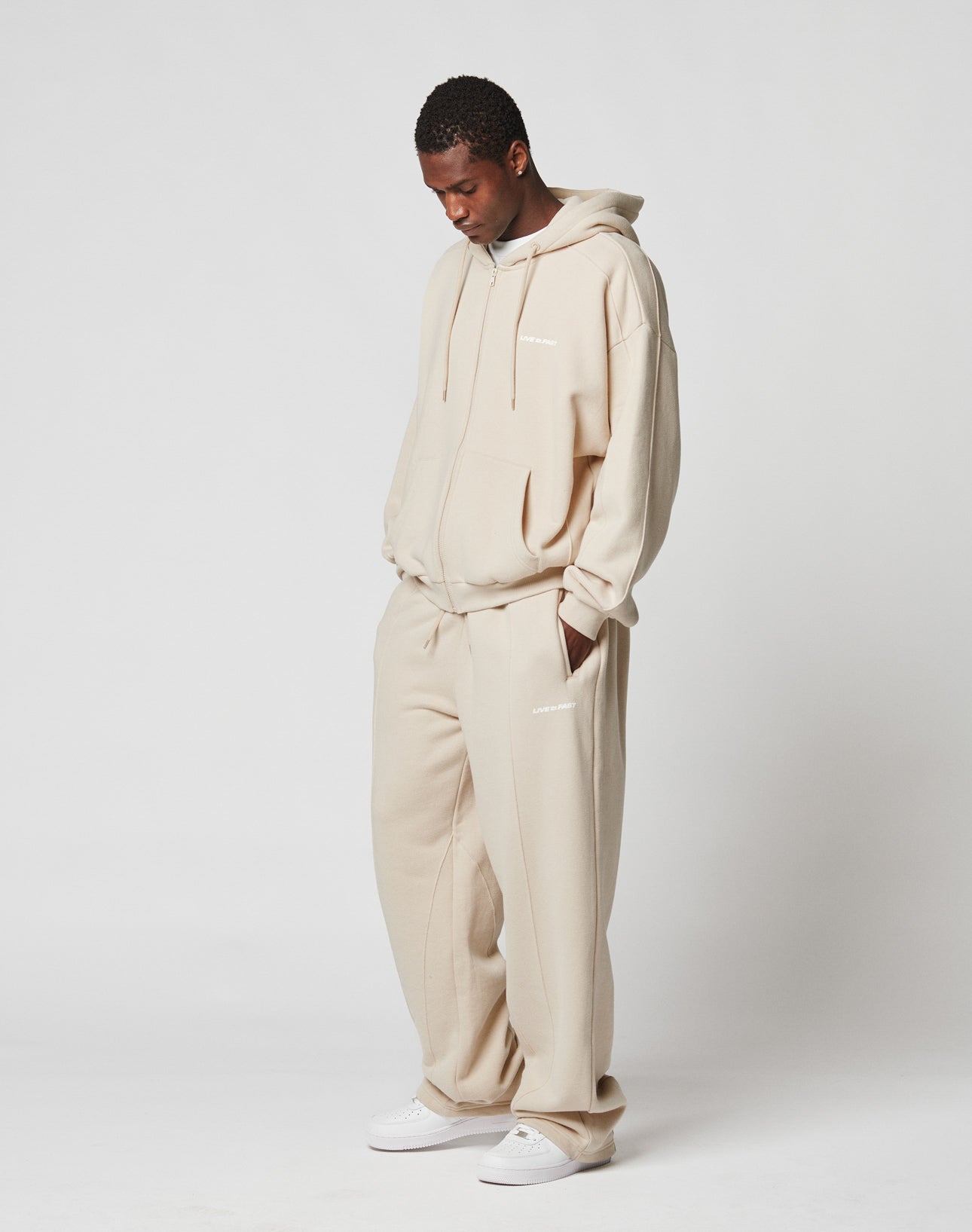 A person stands with head slightly bowed, wearing a wide fit light beige LIVE FAST DIE YOUNG LFDY Pleated Jogger set with white text on both pieces and white sneakers, against a plain white background.