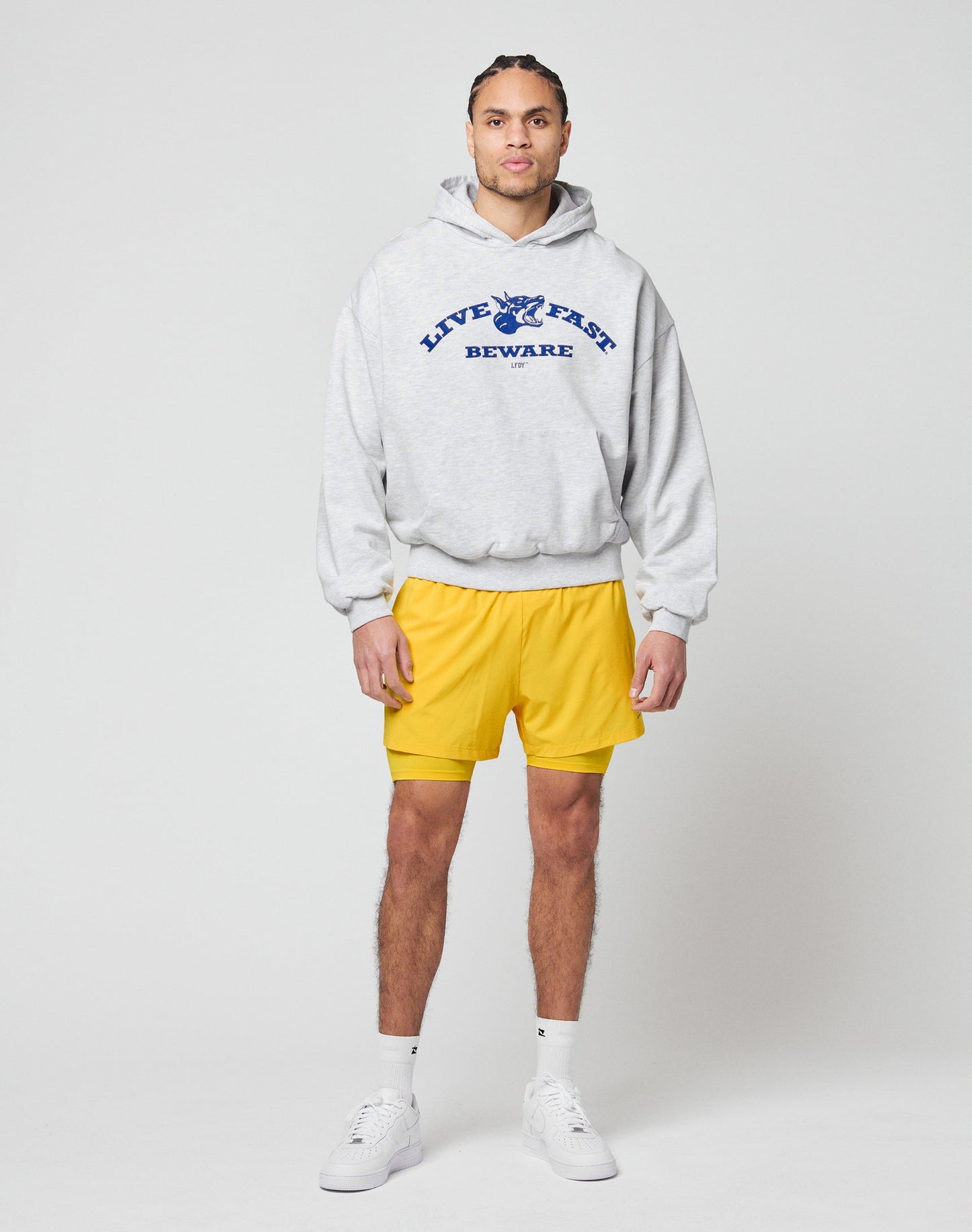 A person stands against a plain background wearing a gray Live Fast Beware Hooded by LIVE FAST DIE YOUNG, featuring a front print, paired with yellow shorts, white socks, and white sneakers.