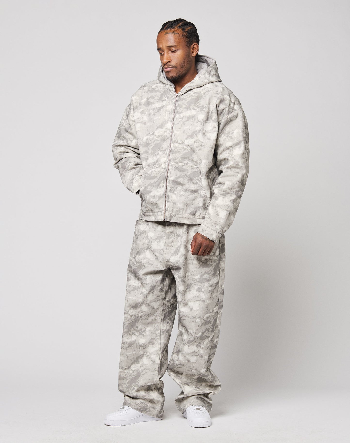 A man stands against a neutral background wearing LIVE FAST DIE YOUNG Live Fast Canvas Pants and a camouflage-patterned hooded jacket, paired with white sneakers. He has his head slightly bowed with one hand in his jacket pocket.