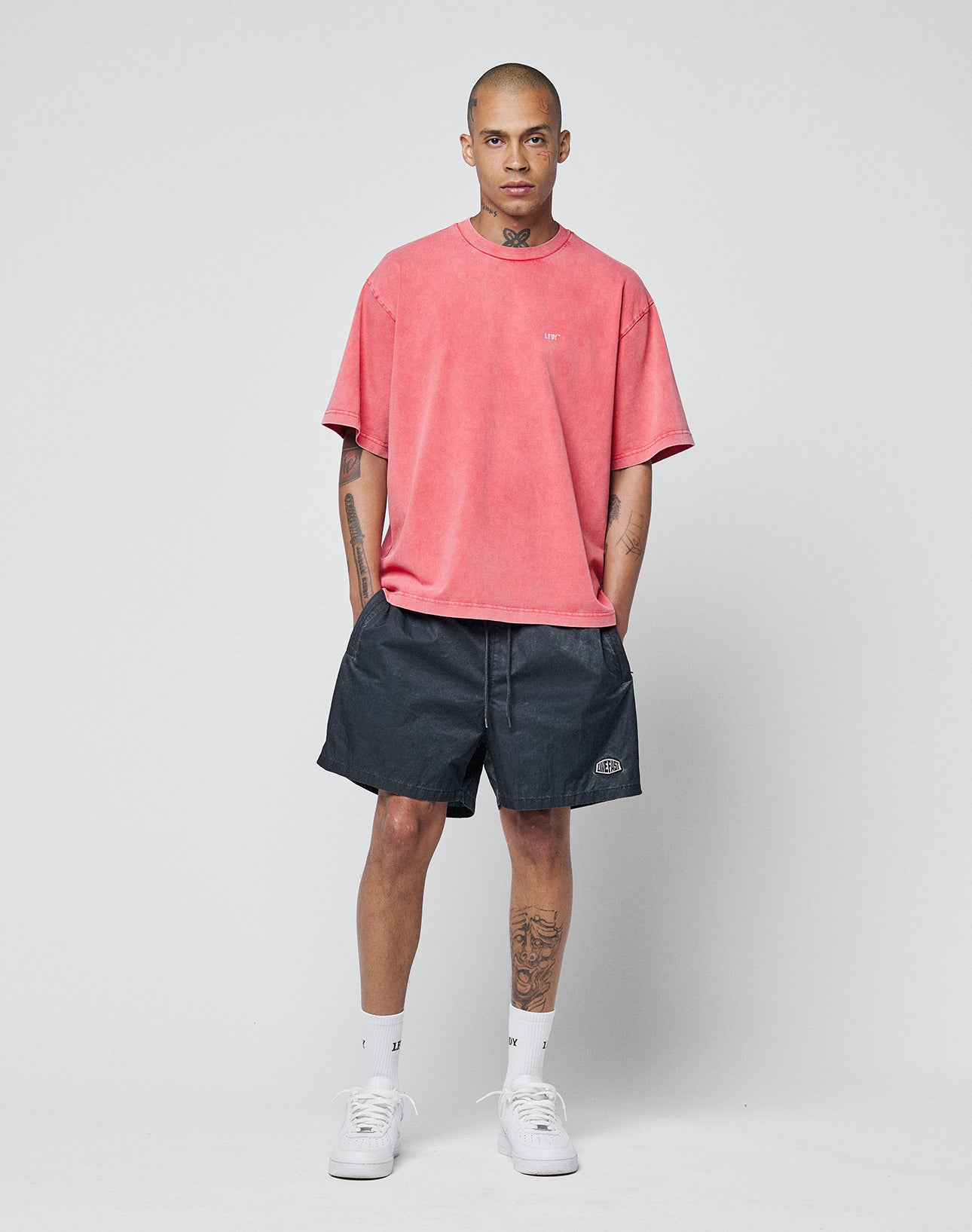 A man with visible tattoos stands against a gray background, wearing a coral LIVE FAST DIE YOUNG Basic Evidence Tee, black shorts, white crew socks and sneakers. His hands are in his pockets and a Flag-Label adds detail to his look.