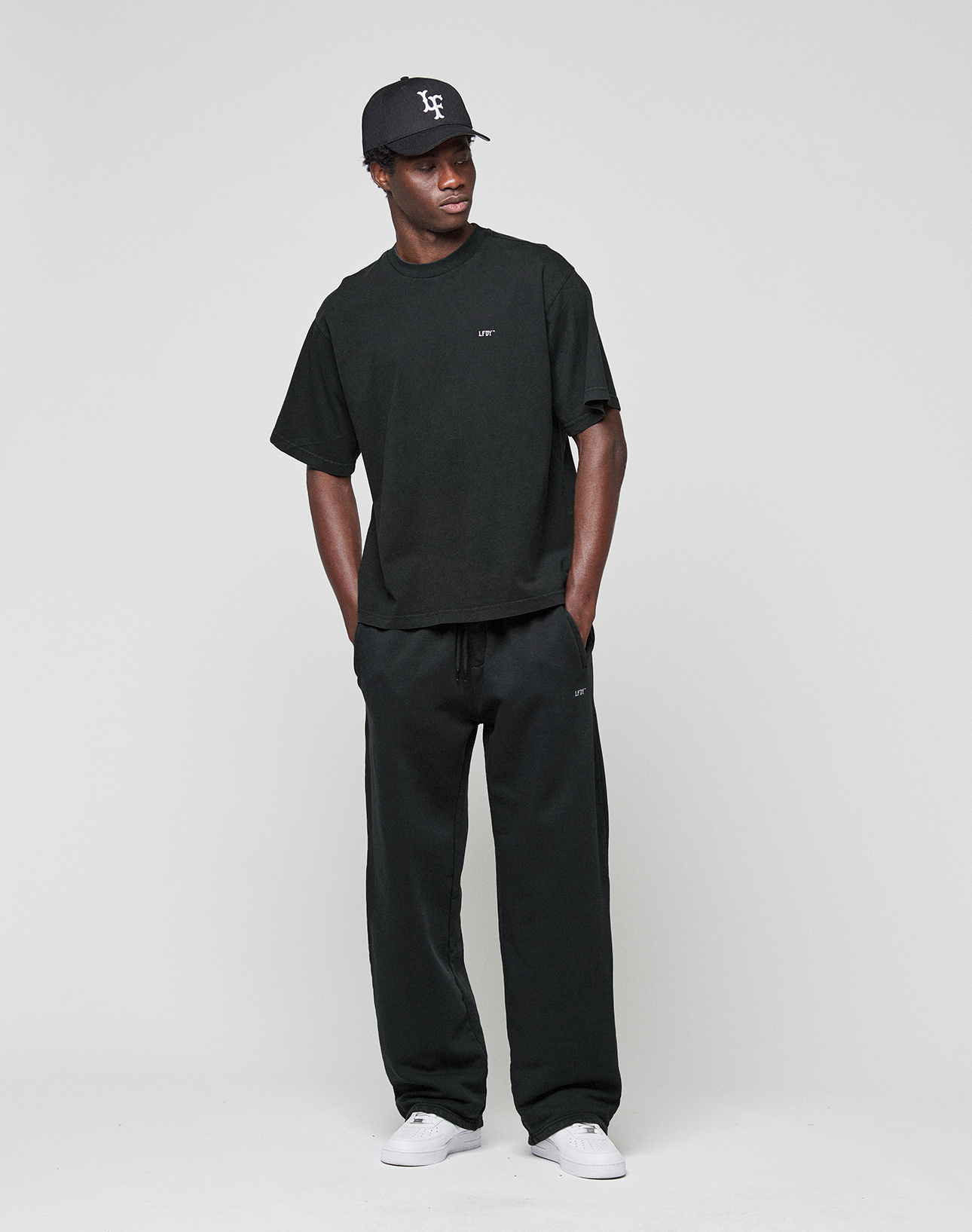 A person poses against a plain background wearing the oversized black 'Basic Evidence Tee' by LIVE FAST DIE YOUNG, paired with black pants, white sneakers, and a black baseball cap. Hands in pockets, they gaze to the side.
