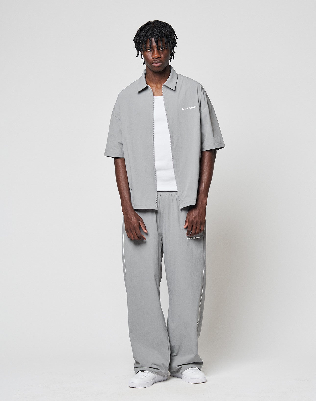 A man with short, curly hair and a relaxed expression wears the LIVE FAST DIE YOUNG LFDY Ripstop Shirt in light gray over a white tee, matched with loose-fitting gray pants and white sneakers against a plain background.