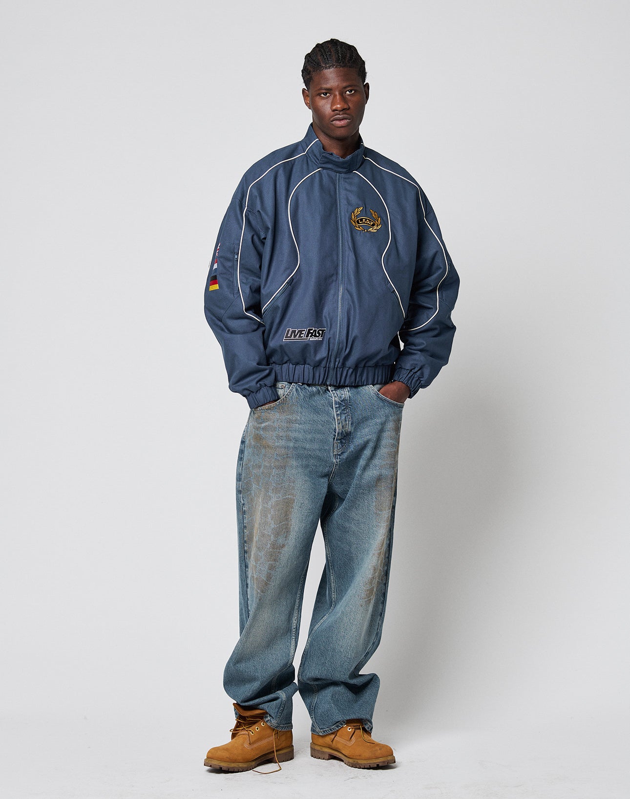A man with short, braided hair poses against a plain backdrop wearing the LFDY Canvas Jacket by LIVE FAST DIE YOUNG, paired with baggy faded jeans and tan work boots. His hands are in his pockets.