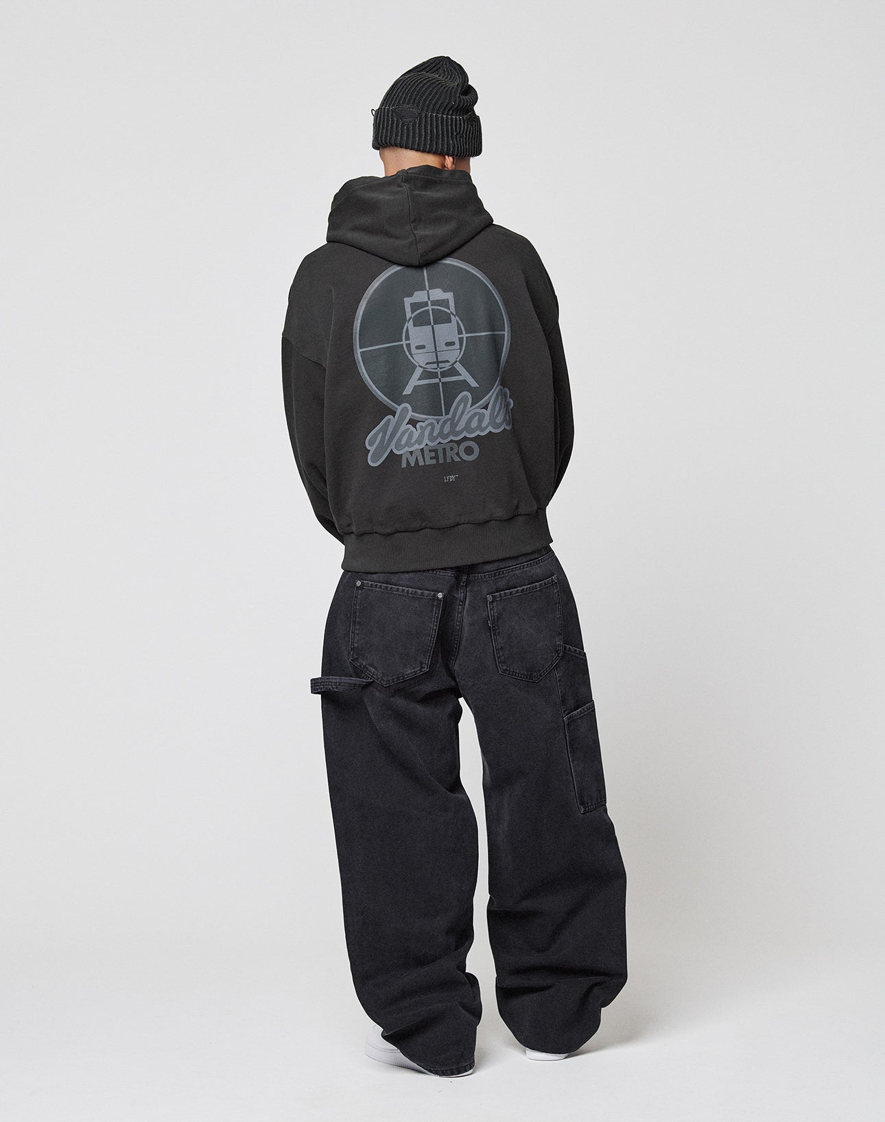 A person faces away, wearing a black beanie, loose black jeans, and the LFDY Vandals Hooded by LIVE FAST DIE YOUNG—an oversize 100% cotton hoodie with a graphic and “Vandal METRO” text on the back, set against a plain light background.