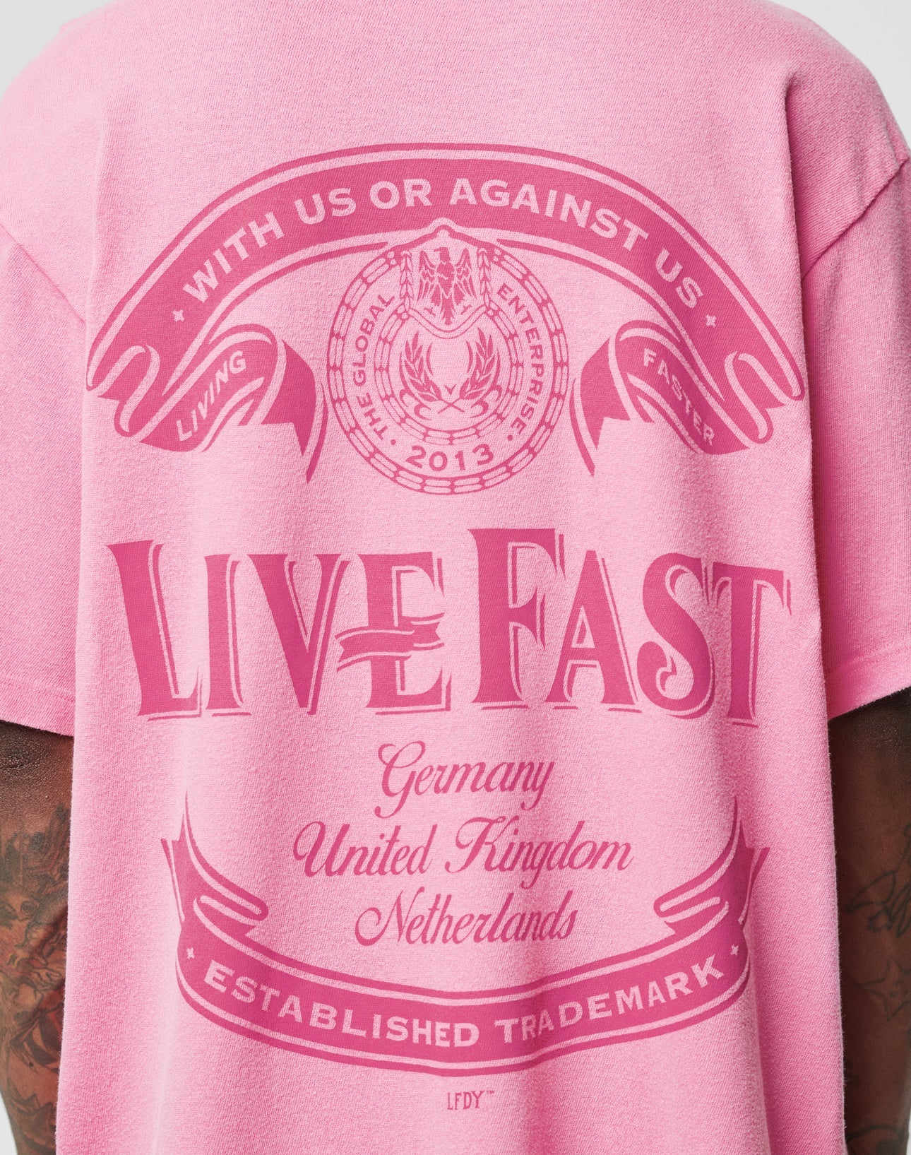 Someone wearing the pink LFDY Loyal Tee by LIVE FAST DIE YOUNG, featuring bold LIVE FAST text on the back, country names (Germany, United Kingdom, Netherlands), banners reading With us or against us and Living Faster. 100% Baumwolle. Oversize Fit.