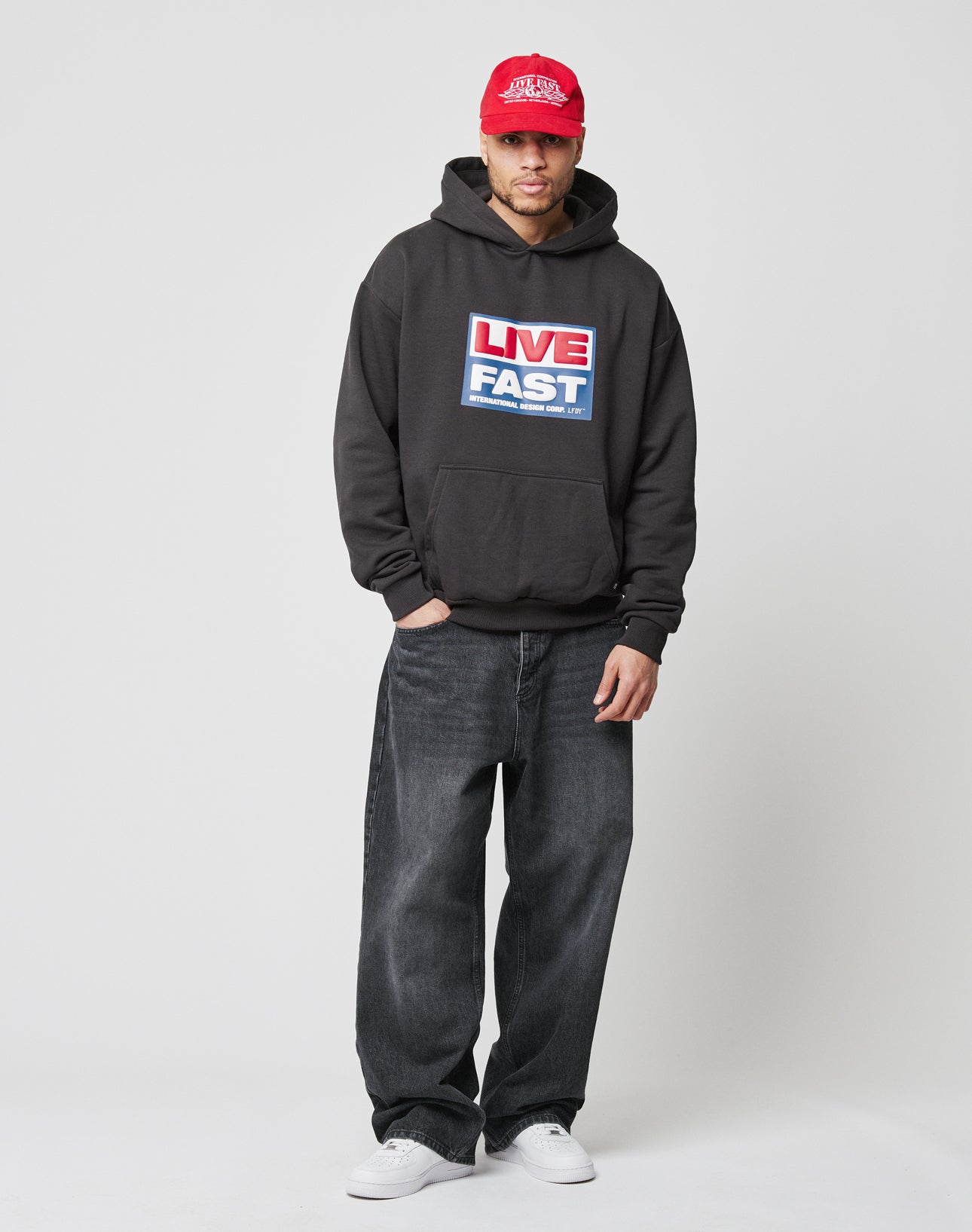 A man stands against a plain background, wearing a red Flag-Label cap, the LIVE FAST DIE YOUNG Live Fast Box Hooded black hoodie with puffer print graphic, oversized baggy black jeans, and white sneakers. His hand is in his pocket and he has a neutral expression.