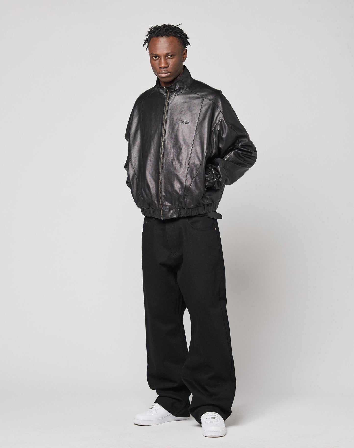 A person stands against a plain background wearing the LIVE FAST DIE YOUNG Live Fast Leather Trackjacket with Stehkragen and embossed logo, paired with loose black pants and white sneakers, hands in pockets and a neutral expression.