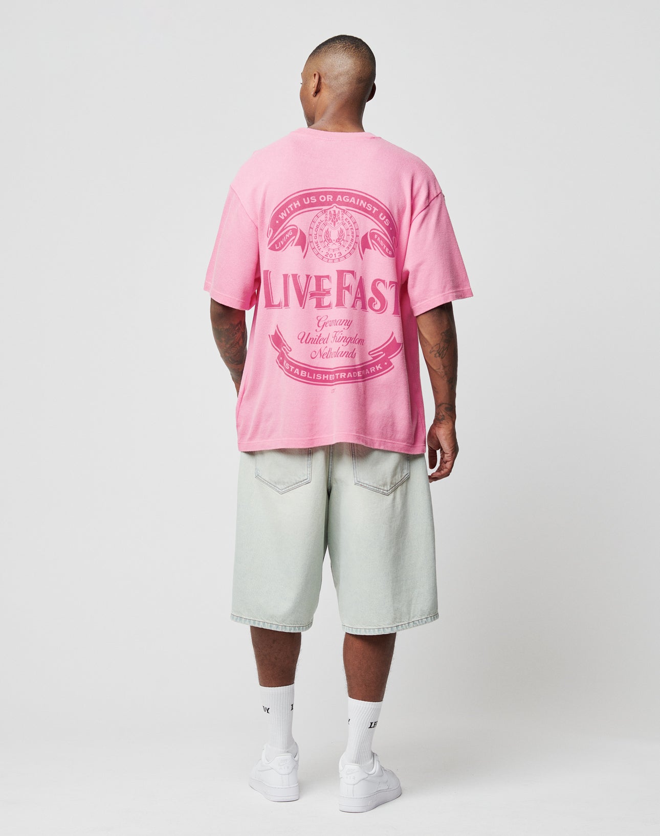 A person faces away, wearing the LIVE FAST DIE YOUNG LFDY Loyal Tee in pink with decorative back text, paired with light washed baggy shorts, white crew socks, and white sneakers against a plain light background.