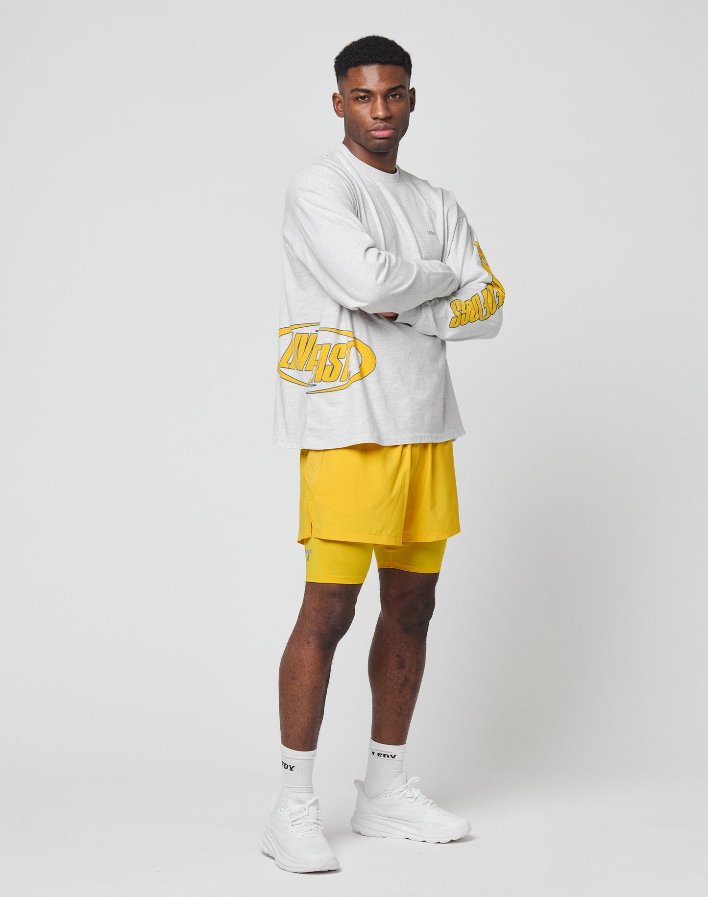 A man stands against a plain white background, confidently looking at the camera with arms crossed. He wears the LIVE FAST DIE YOUNG LFDY Split Sphere Longsleeve in light gray with yellow graphics, paired with yellow shorts, white socks, and sneakers.