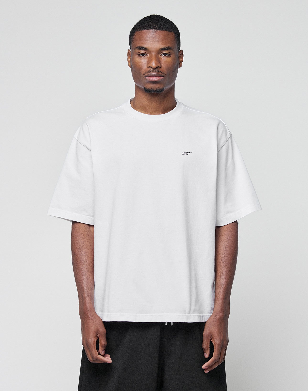 A man faces forward in a LIVE FAST DIE YOUNG Basic Trademark Tee—an oversized, 100% Baumwolle plain white T-shirt with short sleeves and subtle left-chest text—paired with black pants, set against a light gray background.